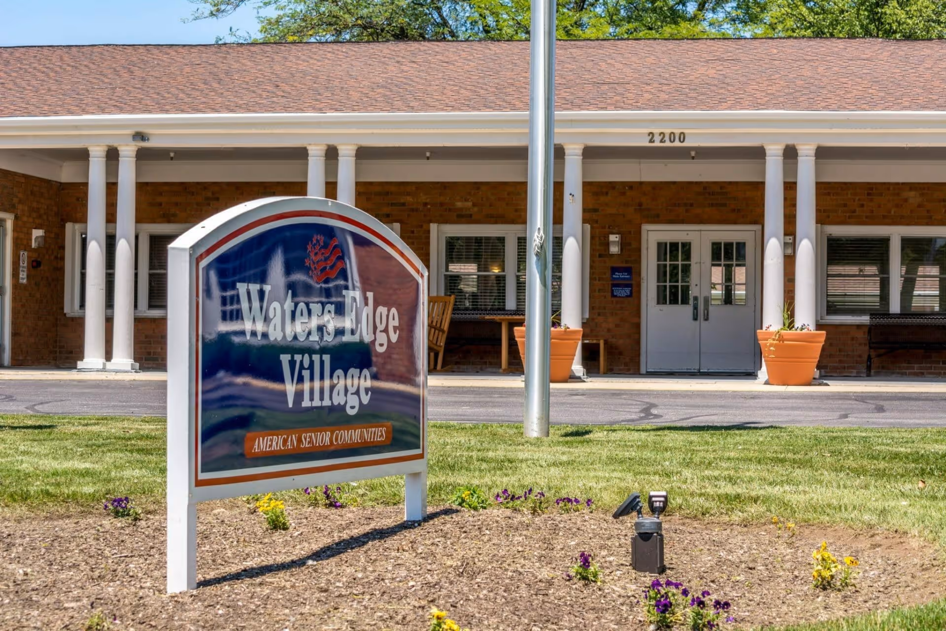 A sign reading "Waters Edge Village" sits in front of the facility entrance with white columns, double doors, and potted plants.