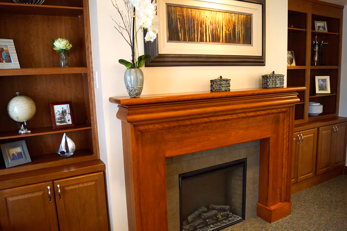 A cozy living room area featuring a wooden fireplace mantel with decorative items on top, flanked by built-in wooden shelves and cabinets displaying framed photos, a globe, a small sailboat sculpture, and vases with flowers.