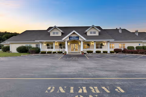 Front exterior view of Jewel Place Senior Living building at dusk with a large parking lot in front and well-maintained landscaping around the entrance.