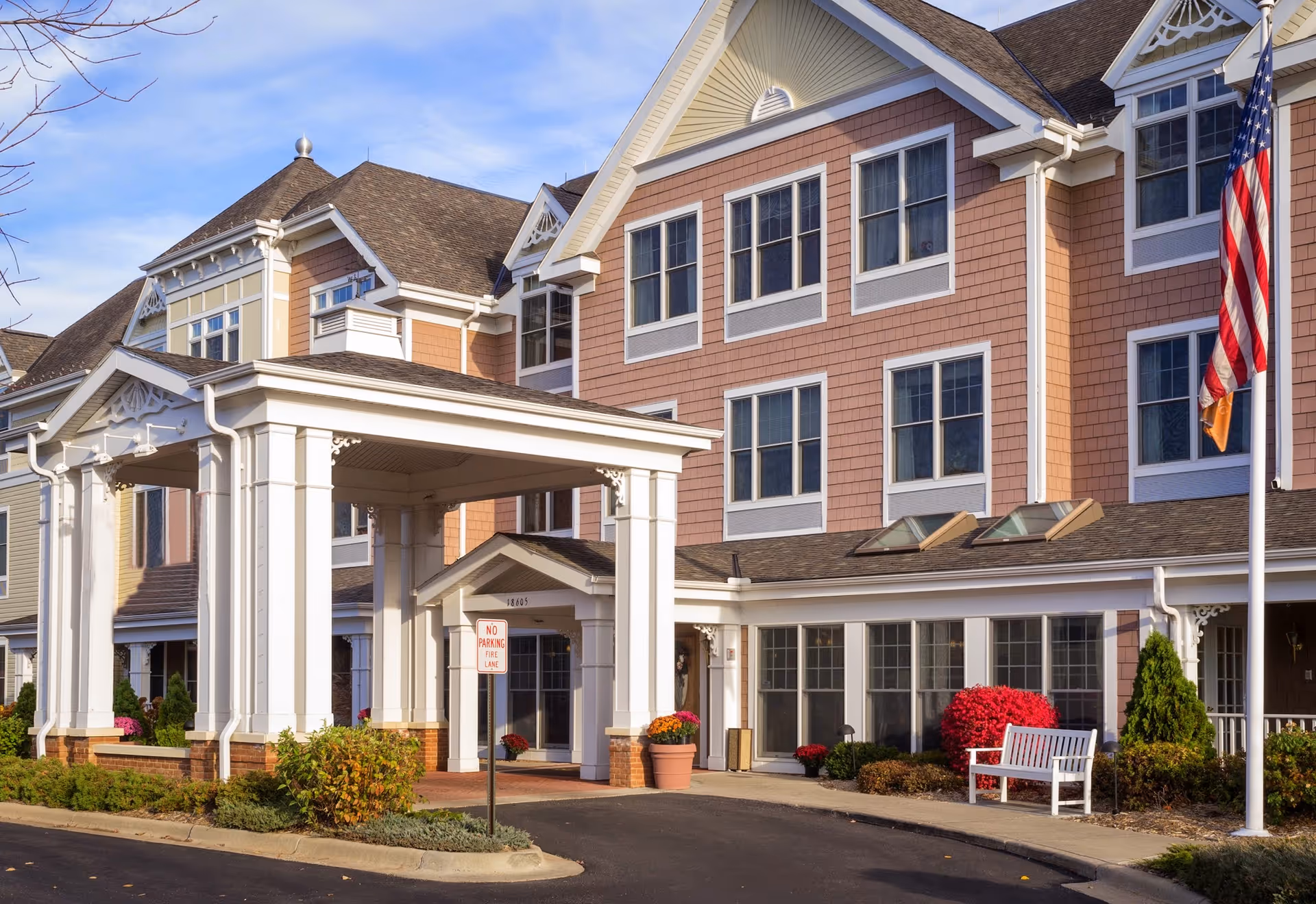 Exterior view of a senior living facility named New Perspective Minnetonka, featuring a large covered entrance with white pillars, multiple windows, a white bench, potted plants, and an American flag on a flagpole.