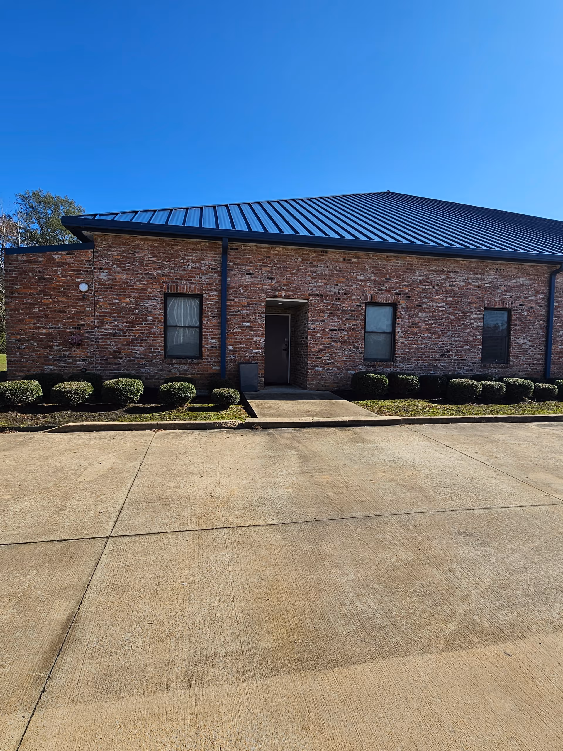 Brick single-story building with a blue metal roof, entry door, and neatly trimmed shrubs.
