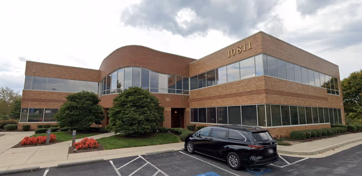 Two-story brick building with curved glass facade, large windows, and a parking lot in front.