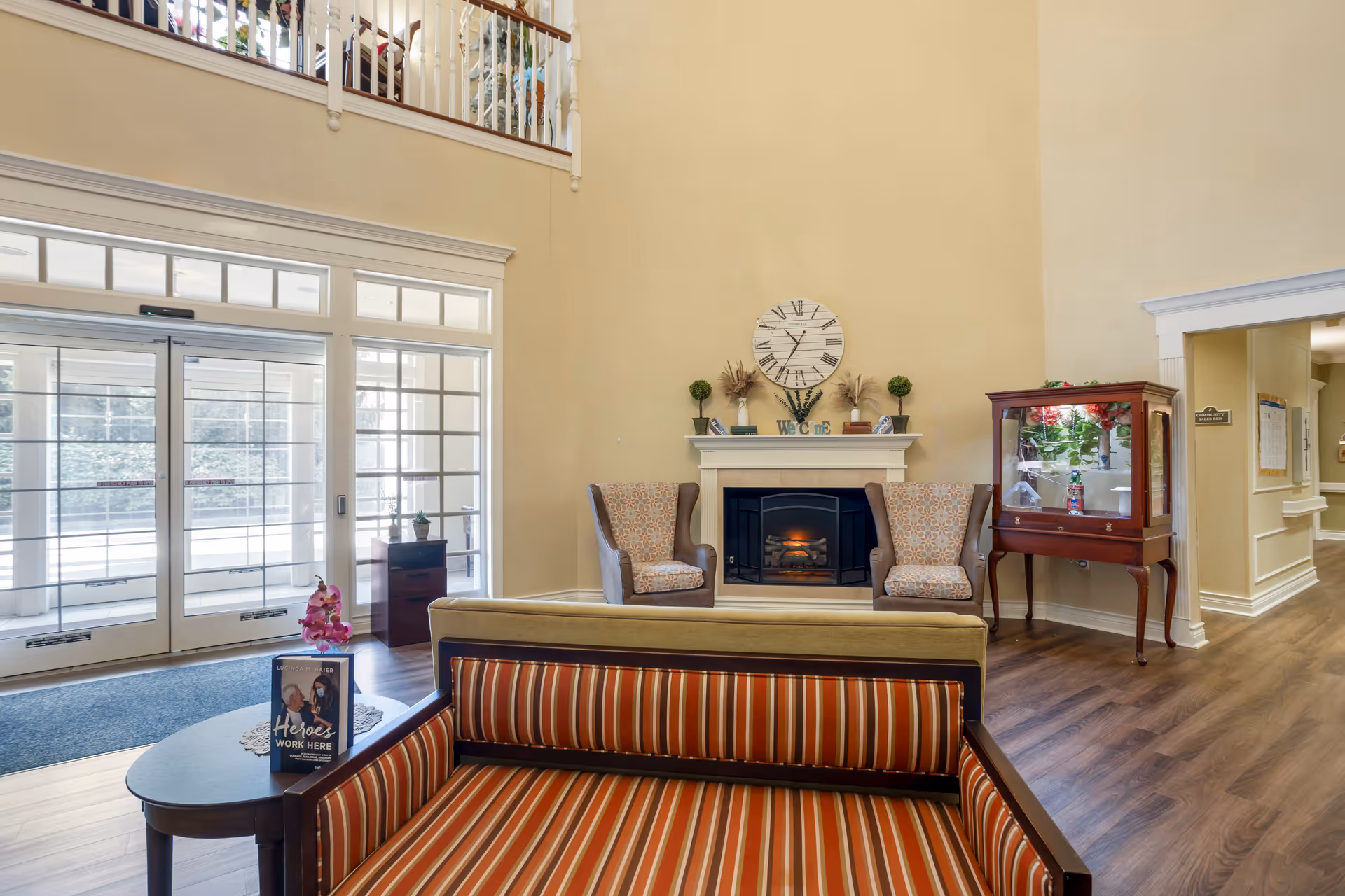 A bright and welcoming senior living facility lobby with a high ceiling, a fireplace flanked by two patterned armchairs, a large clock above the mantel, and a glass display cabinet to the right. In the foreground, there is a striped cushioned bench and a small table with a sign that reads 'Heroes Work Here.' Large glass doors let in natural light from outside.