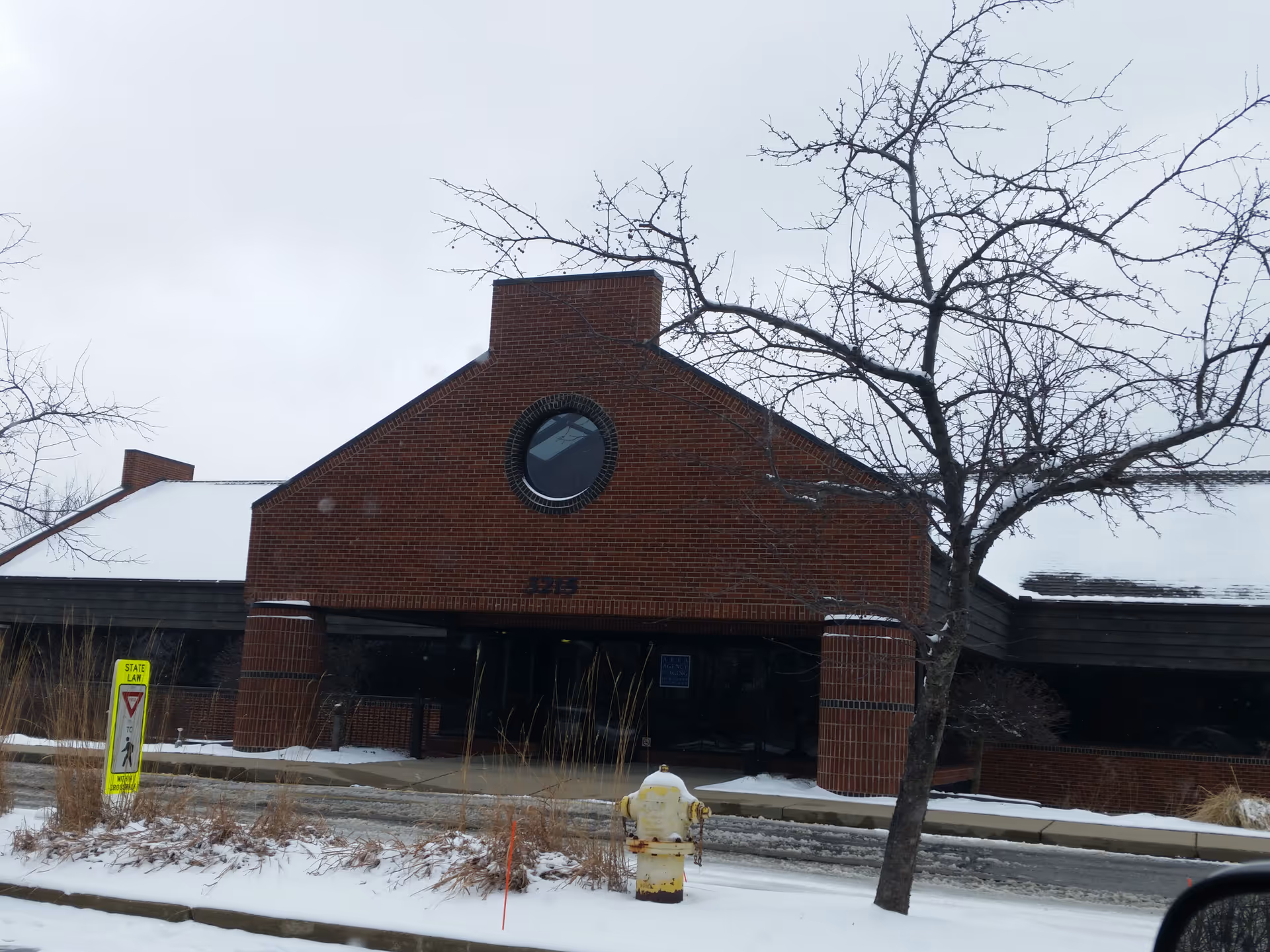 Brick facility front with round window, snow on the ground, and a bare tree.