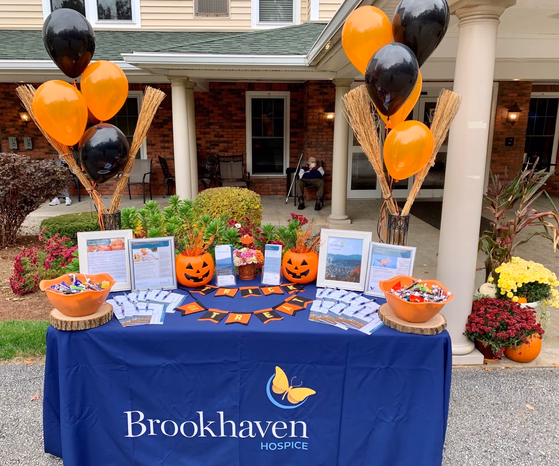 Promotional table for Brookhaven Hospice outside a brick building with orange/black balloons and pumpkins.