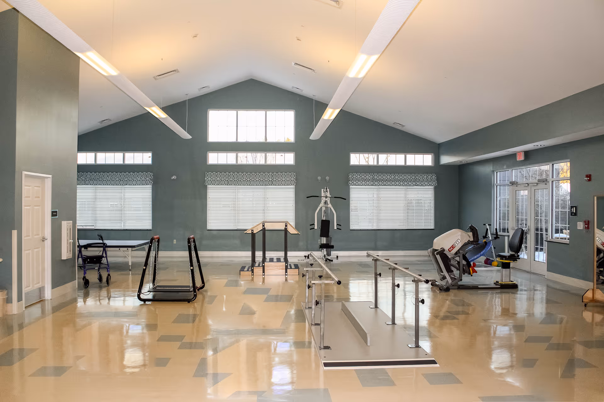 Large bright therapy and exercise room with parallel bars, various exercise equipment, and tall windows under a vaulted ceiling.
