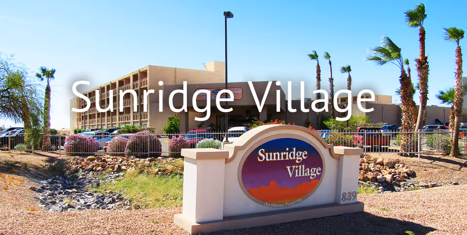 Exterior view of Sunridge Village Retirement facility with a large sign in the foreground displaying the name Sunridge Village. The building is a multi-story structure with balconies, surrounded by palm trees, bushes, and a parking lot with cars. The sky is clear and blue.