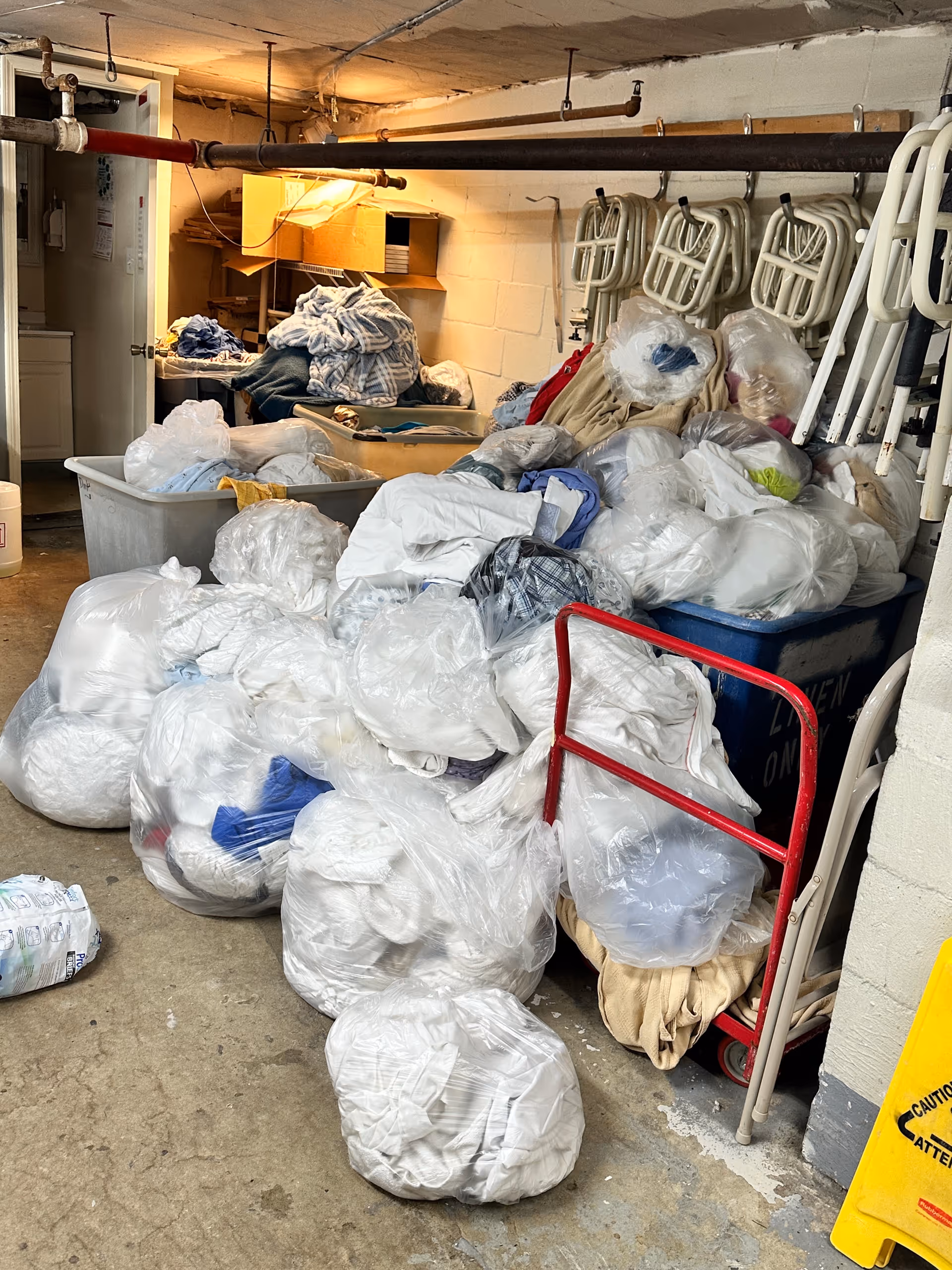 Basement utility room cluttered with laundry bags, bins, and stacked folding chairs.