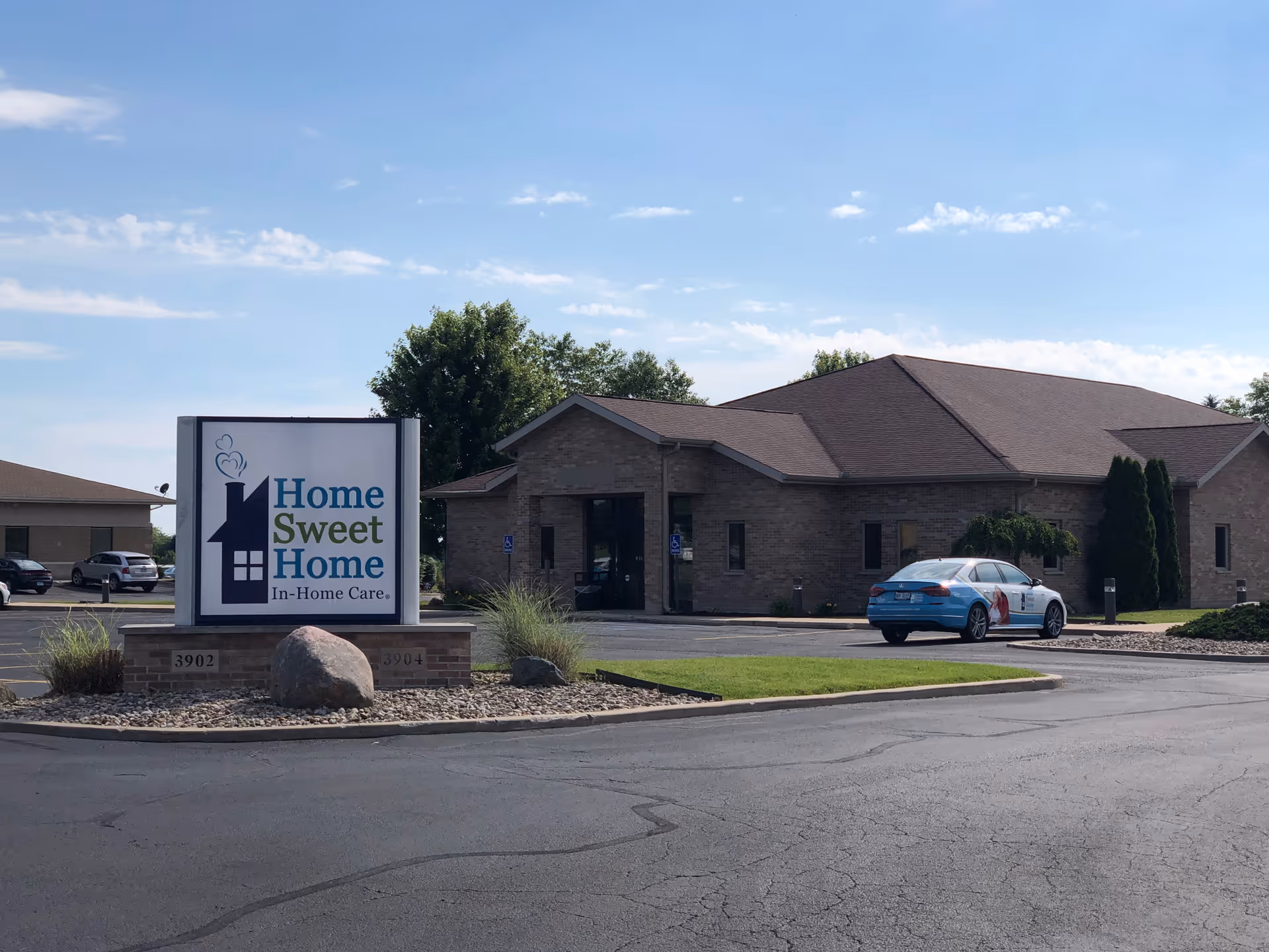 Brick senior living building with sign, parking lot, and blue car outside on a sunny day.