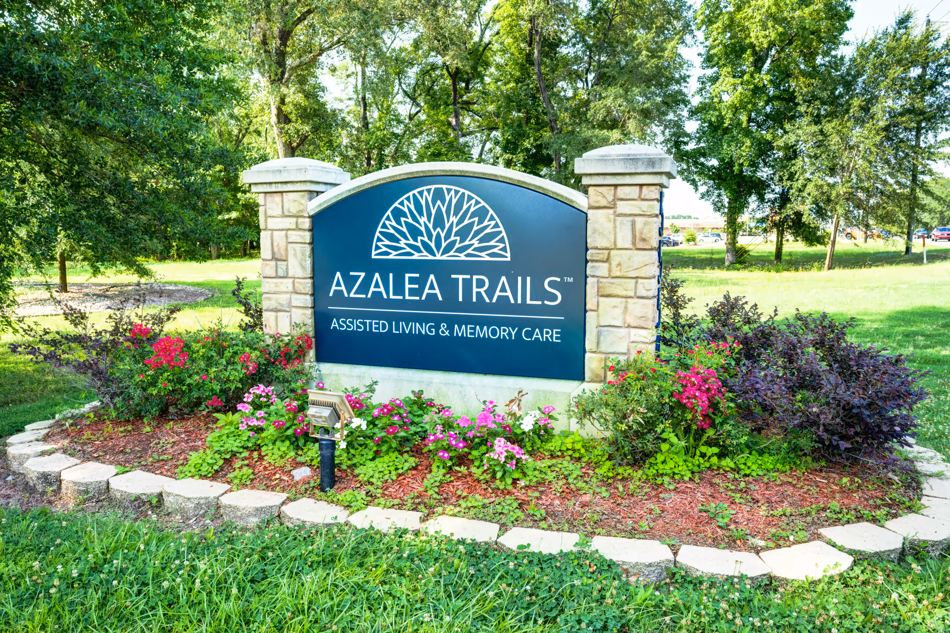 A landscaped stone entrance sign that reads 'AZALEA TRAILS ASSISTED LIVING & MEMORY CARE' surrounded by flowers and trees.