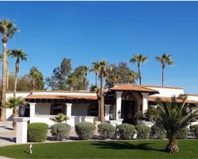 Single-story Mediterranean-style building with tiled roof, columns, palm trees, and a manicured lawn under a clear blue sky.