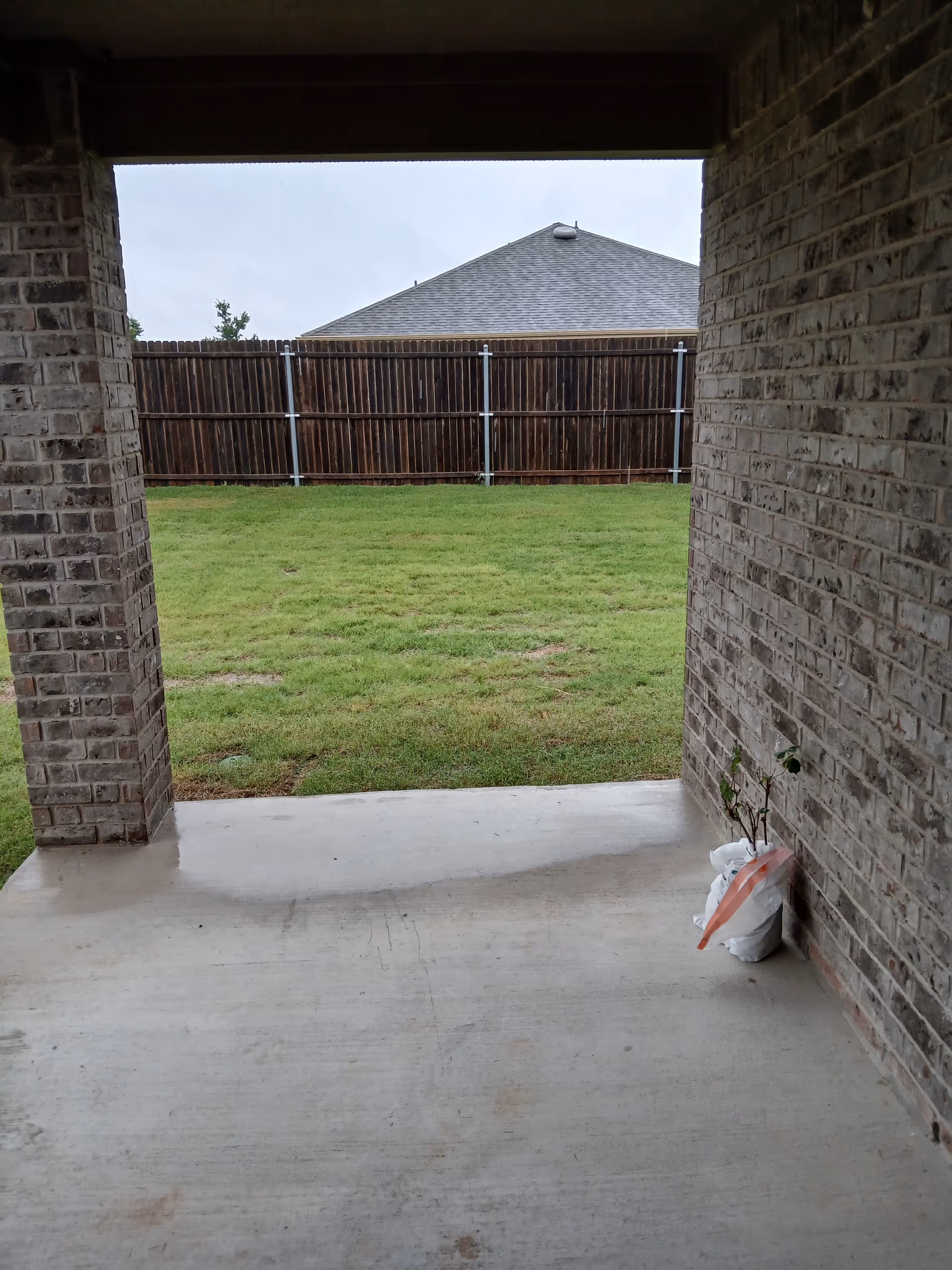Covered brick patio overlooks a grassy yard enclosed by a wooden fence.