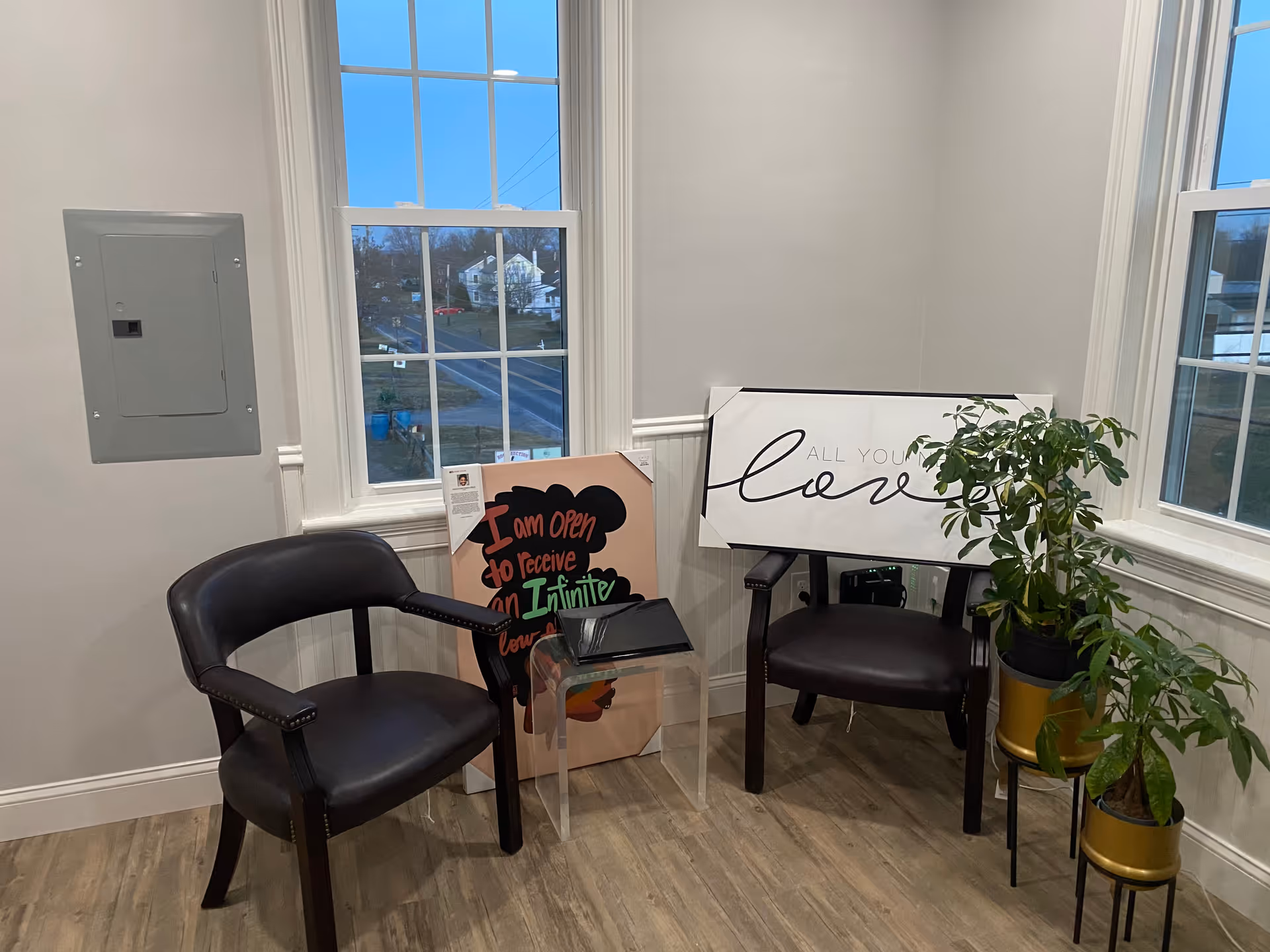 Two black chairs, a small glass table, signs, and a plant near large windows.