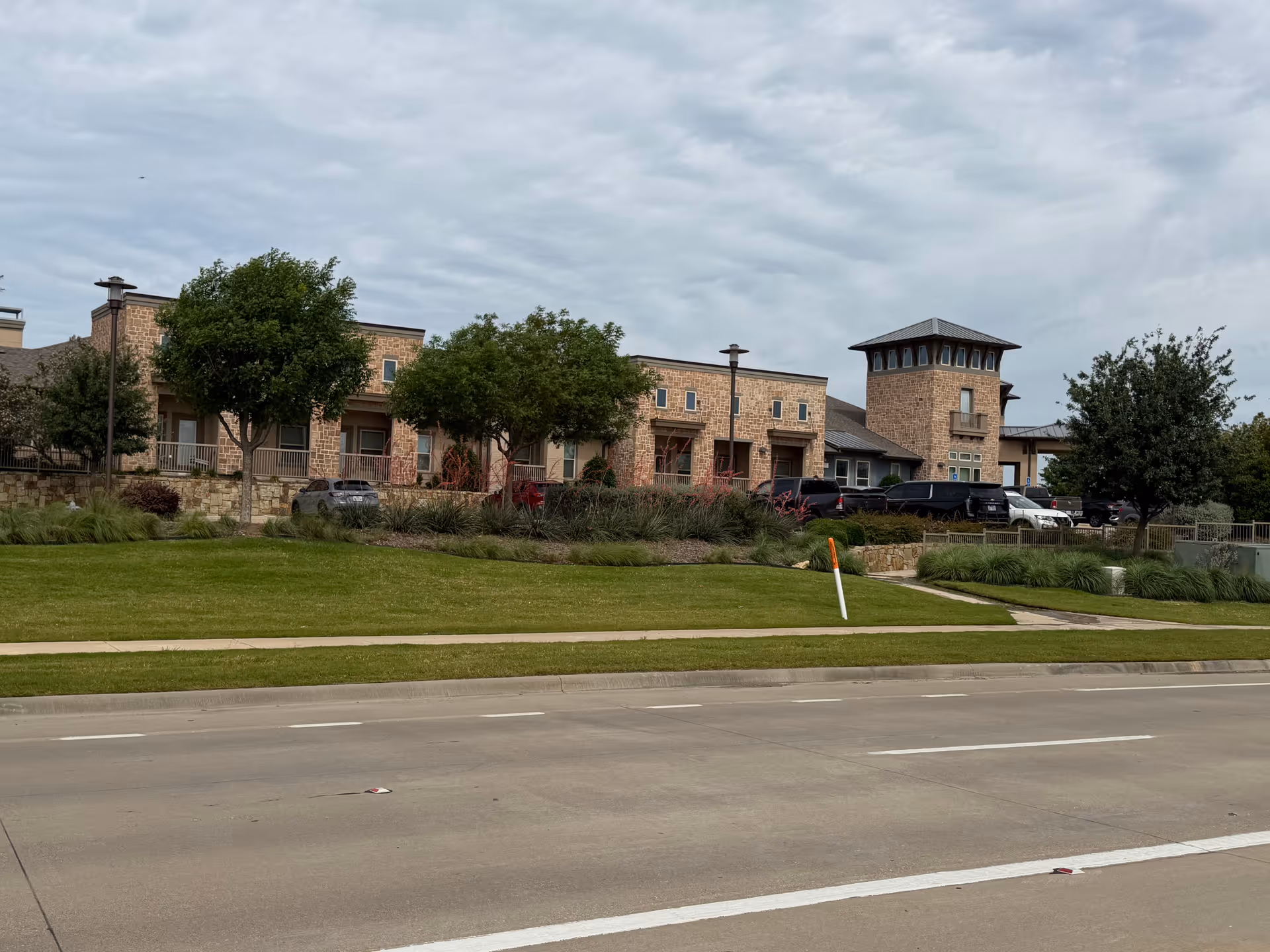 Stone senior living building with a landscaped lawn and parked cars along a street.