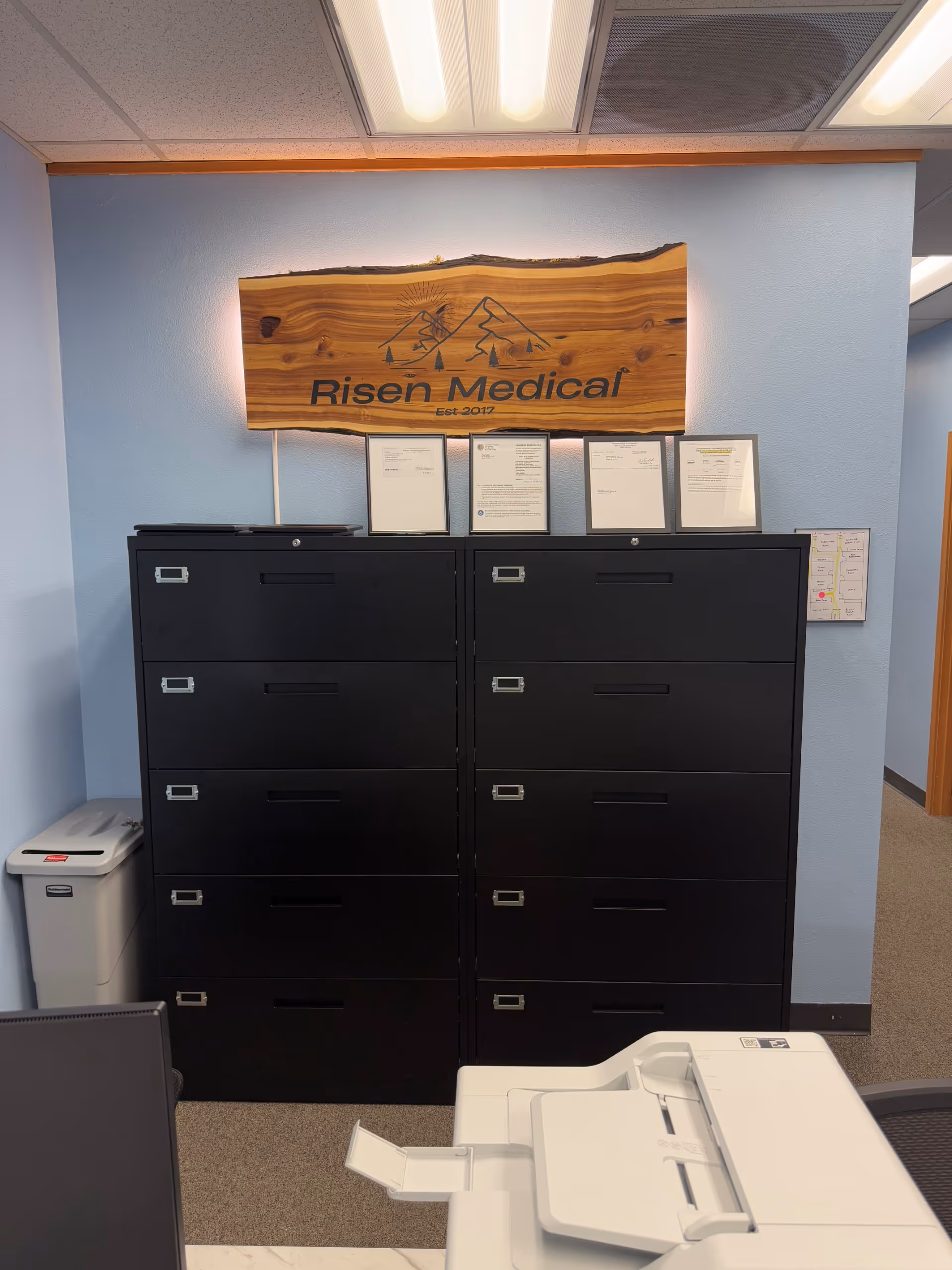Interior office area with filing cabinets, a printer, and a wooden Risen Medical sign.