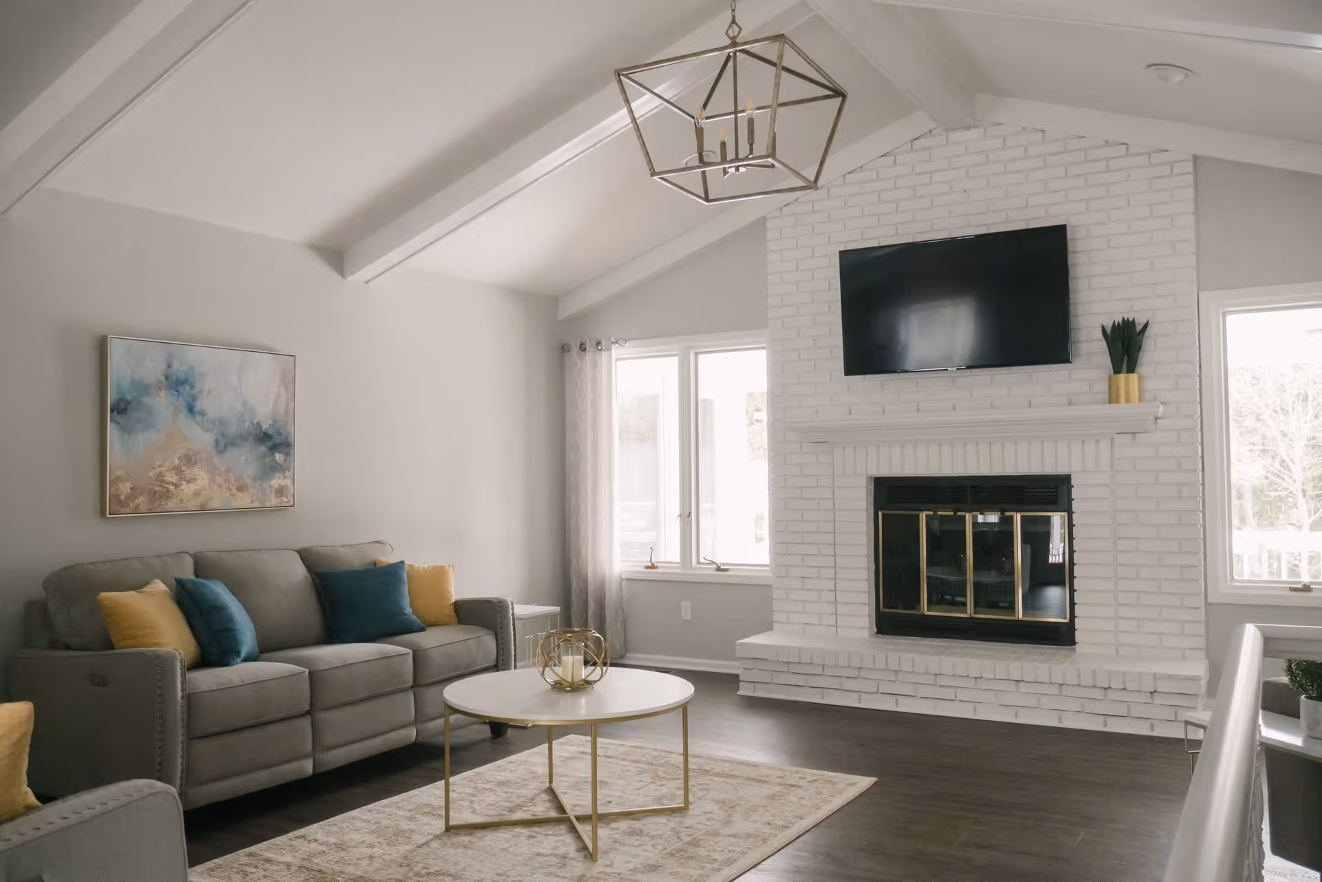 A bright living room with a vaulted ceiling featuring exposed beams, a gray sofa with blue and yellow cushions, a round white coffee table with a decorative candle holder, a white brick fireplace with a mounted flat-screen TV above it, and two windows with sheer curtains letting in natural light.