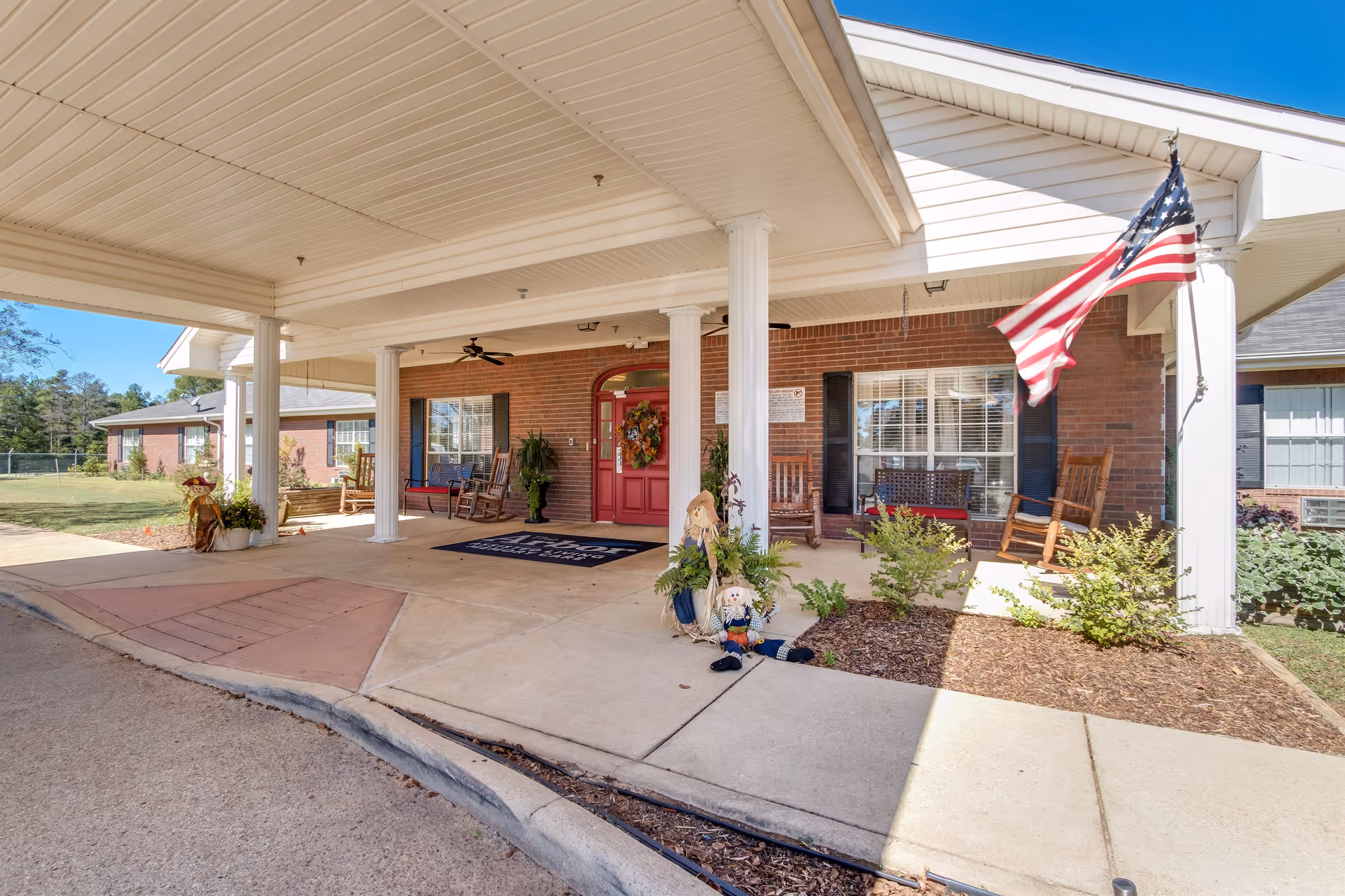 Covered entrance of a brick building with white columns, a red door decorated with a wreath, rocking chairs on the porch, potted plants, and an American flag hanging from one of the columns. There are scarecrow decorations near the entrance and a welcome mat on the ground.