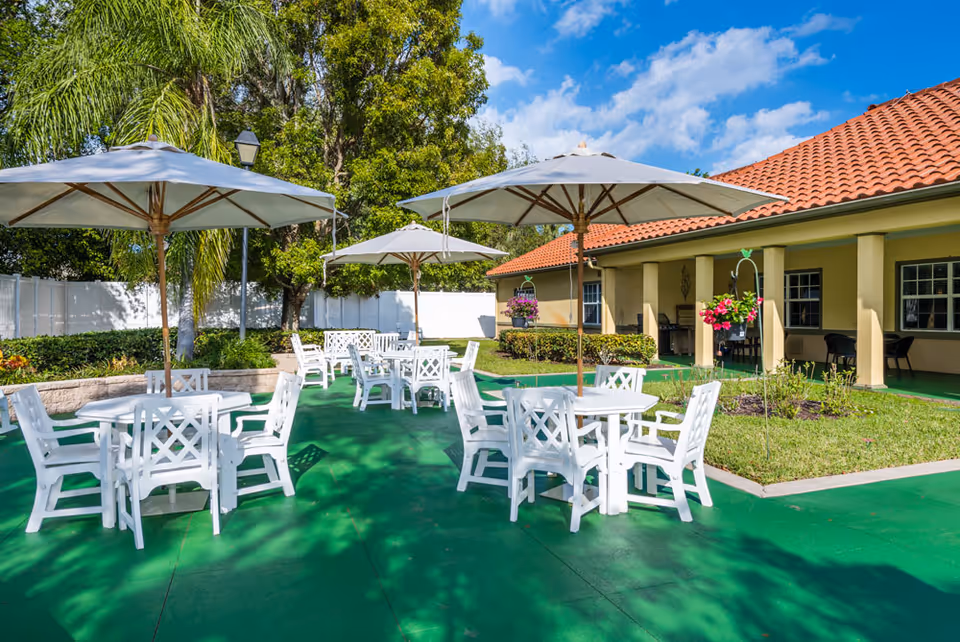 Outdoor patio area with white tables and chairs under large white umbrellas. The patio has a green floor and is surrounded by trees, bushes, and a building with a red tiled roof. Hanging flower baskets add color to the scene under a partly cloudy blue sky.