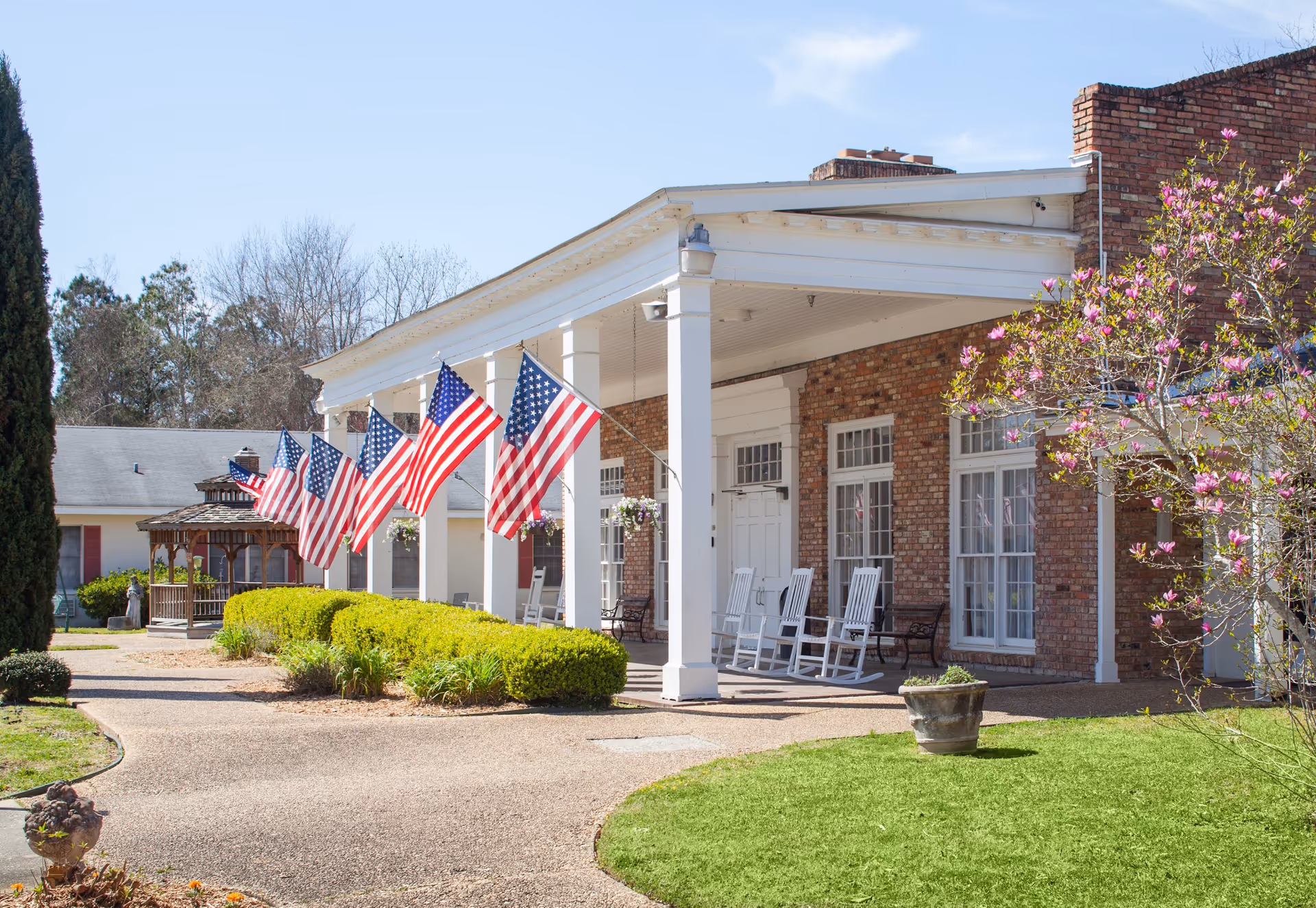 Brick senior living facility front porch with rocking chairs, American flags, and manicured lawn.