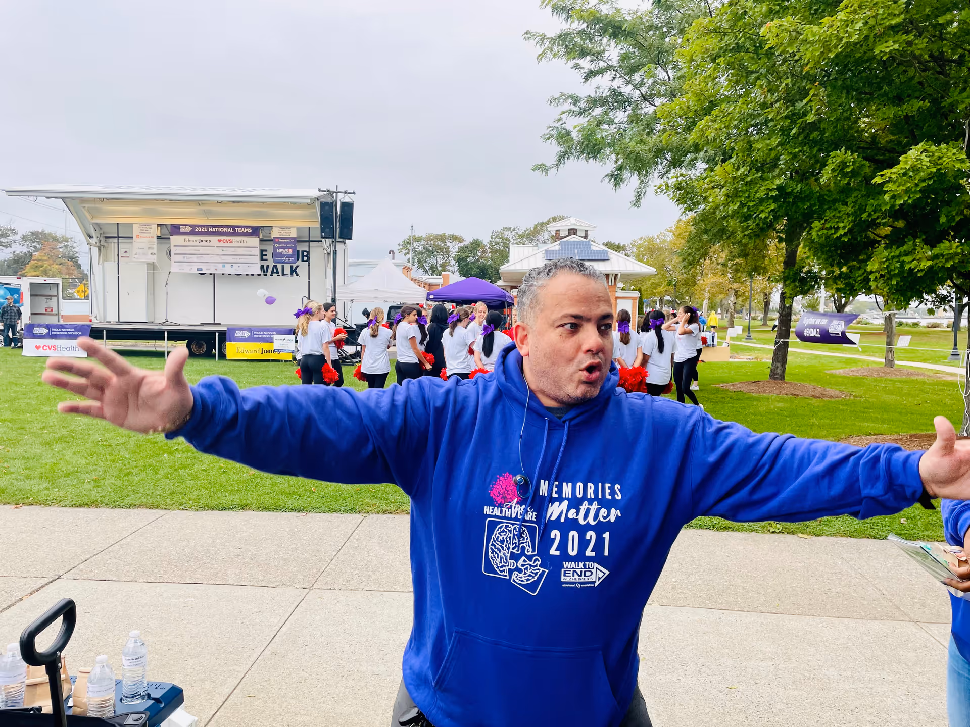 Man in blue hoodie extends arms outdoors at a park event with a group in background.