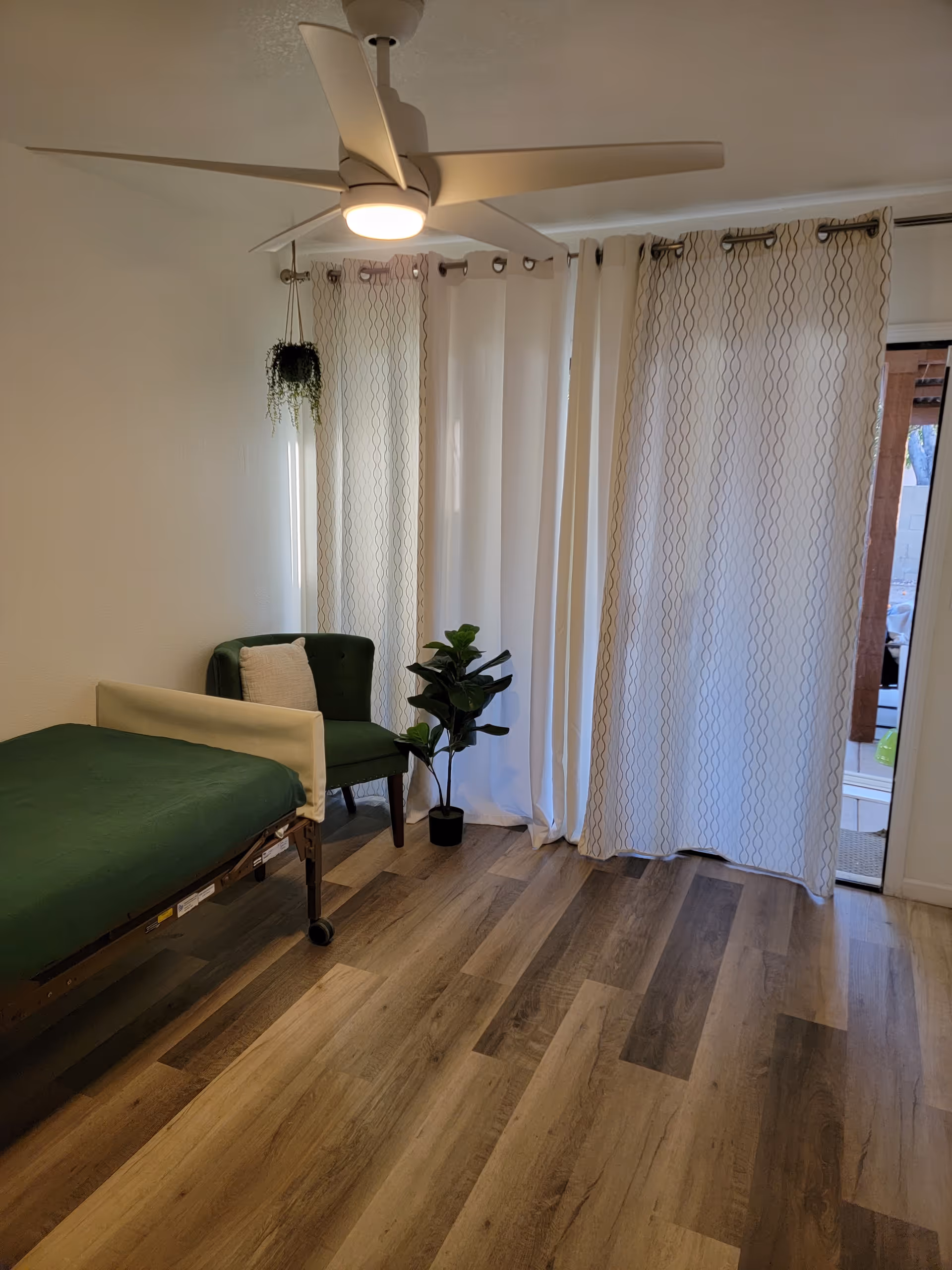 Hospital-style bed with green sheets, a chair, plants, and white curtains in a bright bedroom.