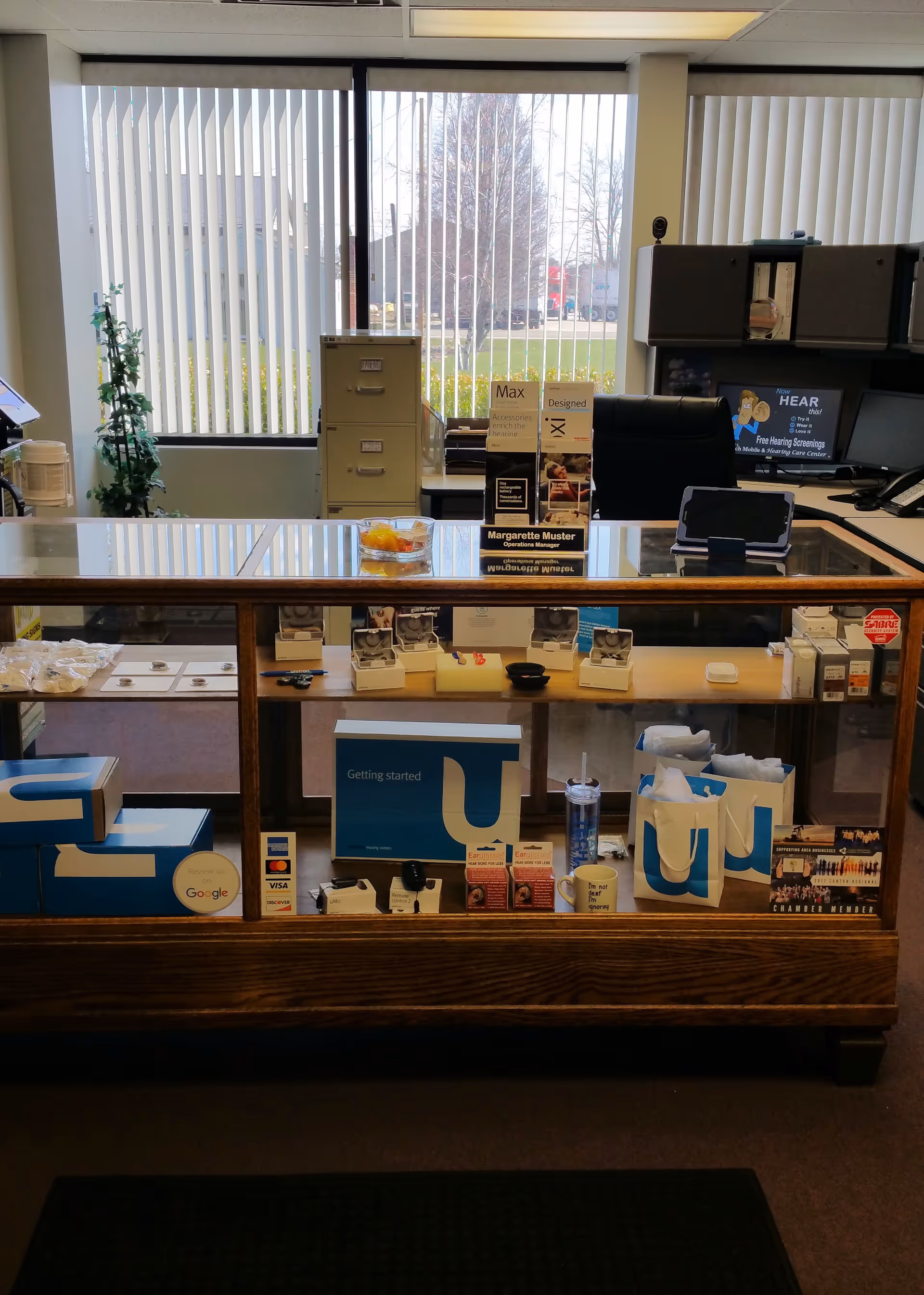 Indoor reception desk with glass display, brochures, and office equipment opposite large vertical blinds by windows.