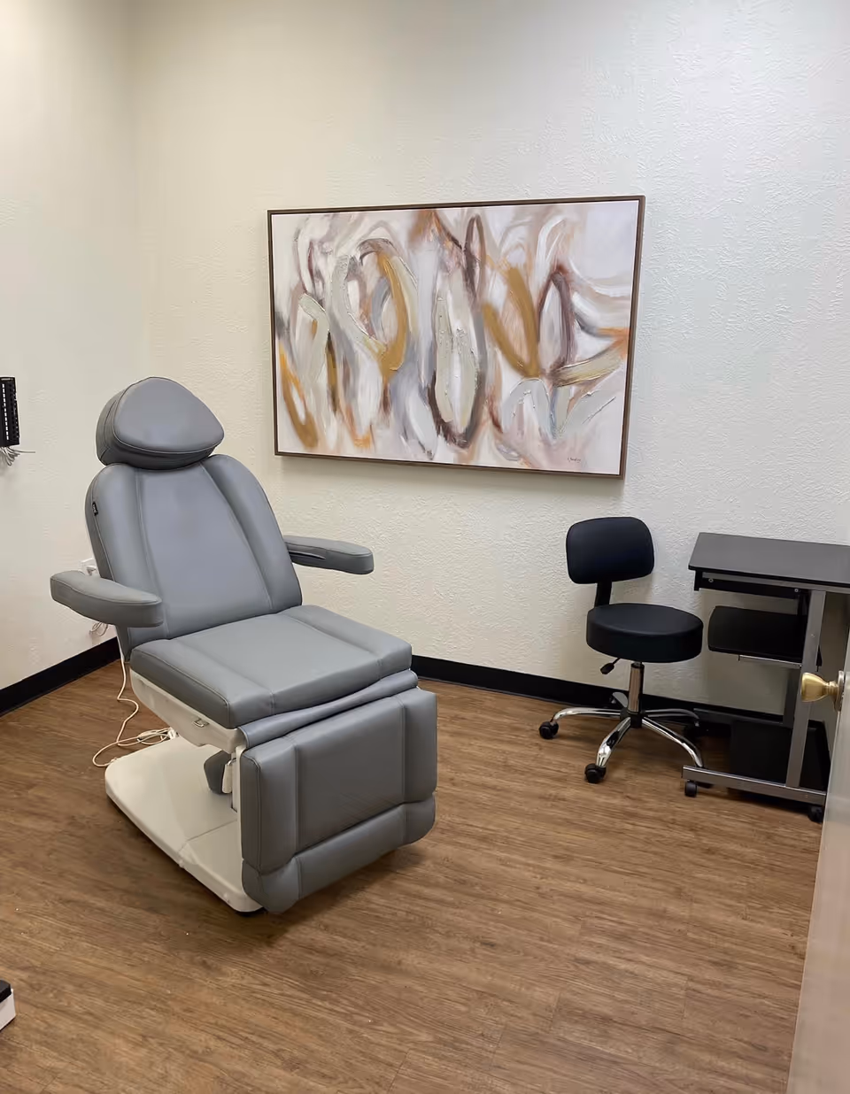 A gray exam chair in a small room with abstract wall art and a black stool.