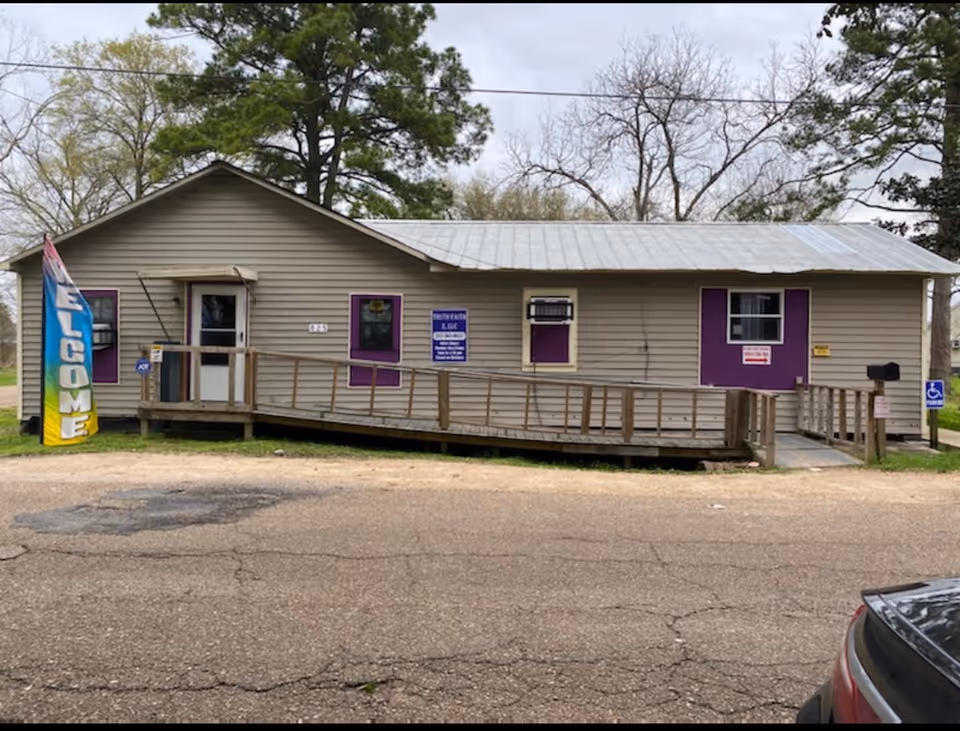 Small beige building with purple accents, wheelchair ramp, flag, and parking in front.