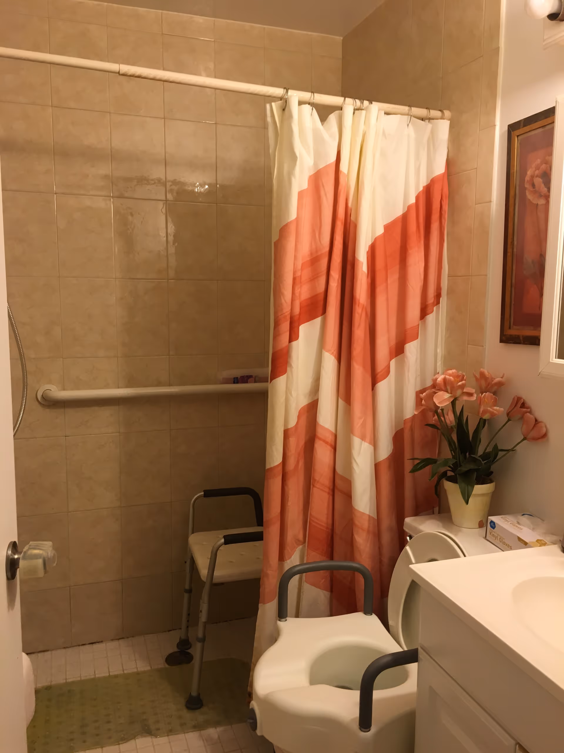 A bathroom with beige tiled walls and a shower area with a grab bar and a shower chair. There is a shower curtain with a red and white pattern partially closed. In front of the toilet is a raised toilet seat with armrests. A white vanity with a sink is visible on the right side, along with a potted plant with pink flowers and a framed floral picture on the wall.