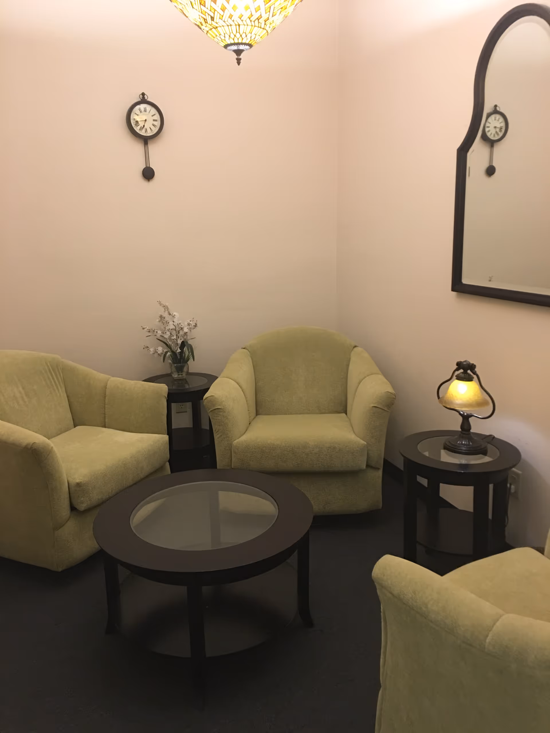 A cozy lounge area with green armchairs, glass-top tables, lamps, and a wall mirror.