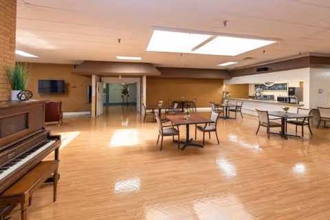 Large multipurpose dining room with hardwood floors, several small tables and chairs, a piano on the left, and a serving counter on the right under skylights.