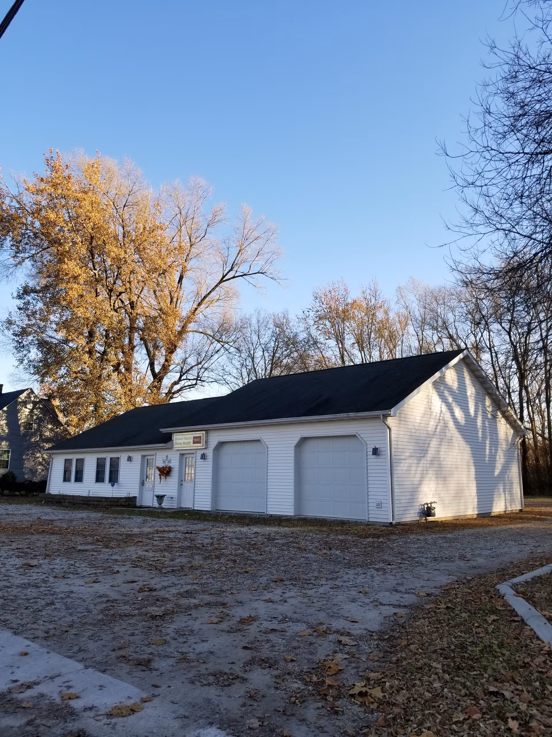 White single-story building with three garage bays sits on a gravel lot among leafless trees.