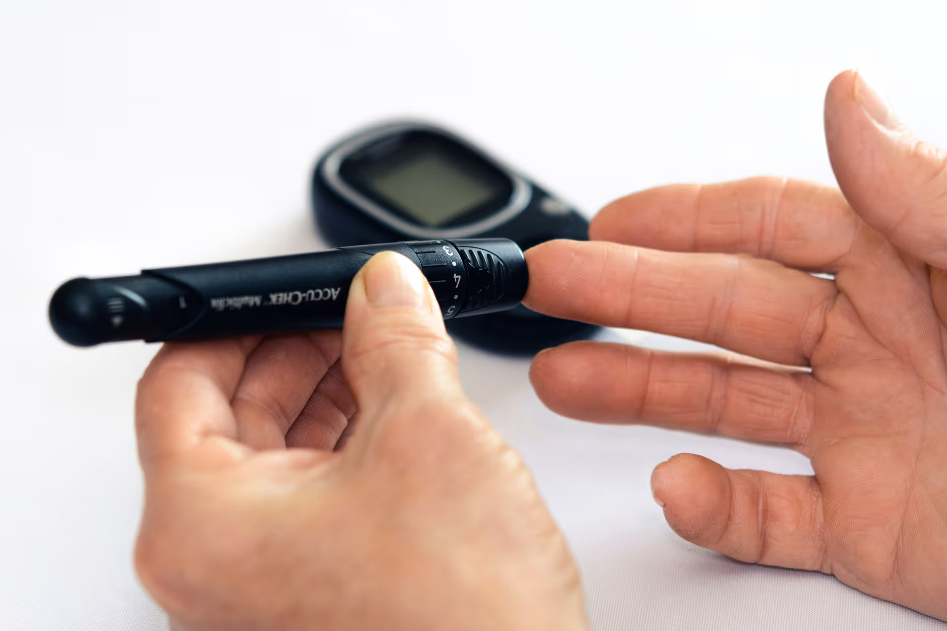 Close-up of hands using a lancet device near a glucometer on a white surface
