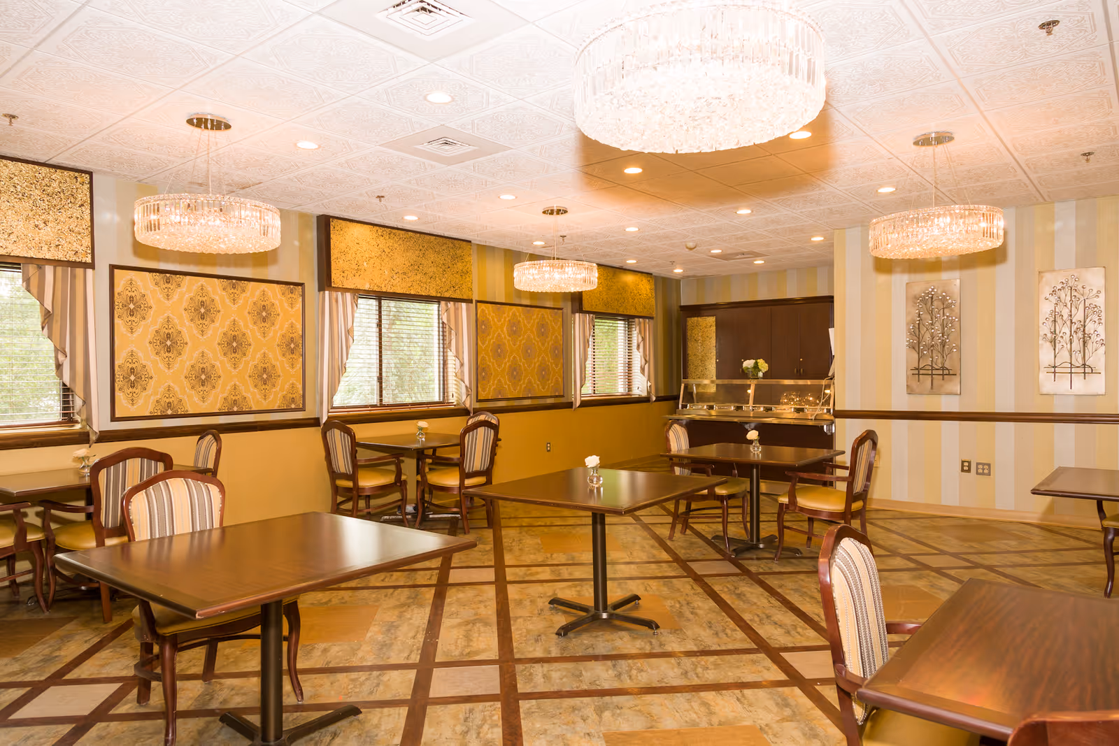 A well-lit dining room with several wooden tables and chairs arranged neatly. The room features large windows with decorative curtains and patterned wall panels. Elegant chandeliers hang from the ceiling, and there are two framed metal art pieces on one wall. The floor has a geometric tile pattern.