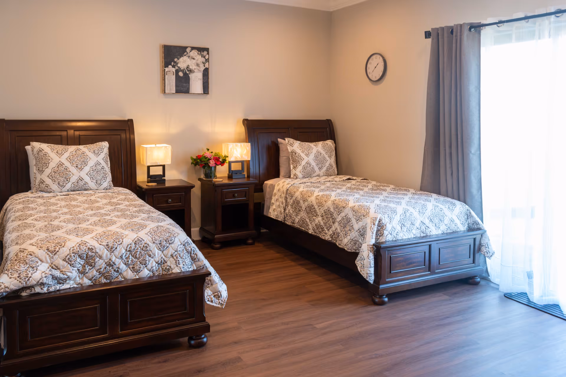 A bedroom with two twin beds featuring dark wooden frames and matching patterned bedspreads. Between the beds are two nightstands with lamps and a vase of flowers. A clock is mounted on the wall, and a window with gray curtains lets in natural light.