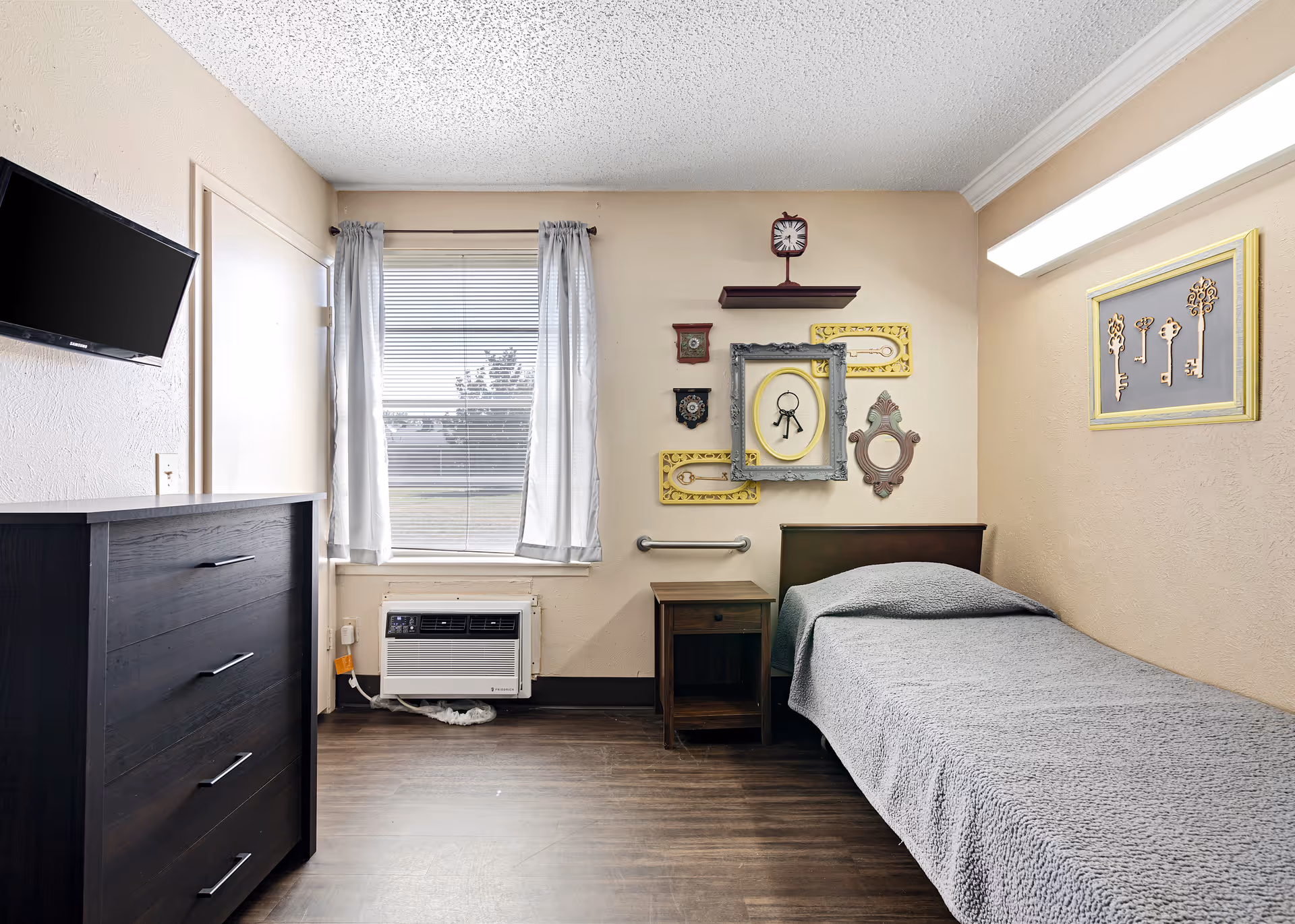 Small nursing-home bedroom with a single bed, nightstand, dresser, mounted TV, window and decorative wall art.