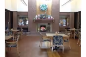 Indoor senior living common room with brick fireplace, shelves, and tables with cushioned chairs.