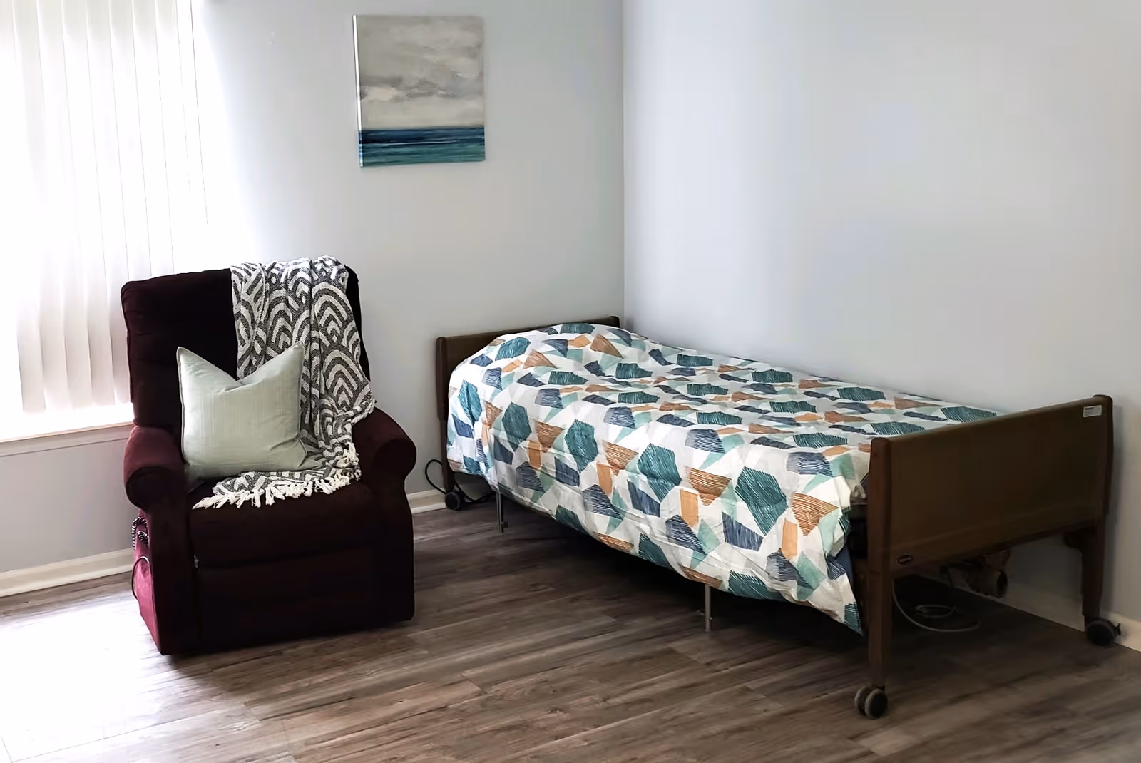 A simple bedroom with a single bed covered with a geometric patterned quilt in shades of blue, green, and brown. Next to the bed is a dark red recliner chair with a light green pillow and a black and white patterned throw blanket. The room has light-colored walls and wood flooring, with a window covered by vertical blinds and a small abstract painting on the wall above the bed.