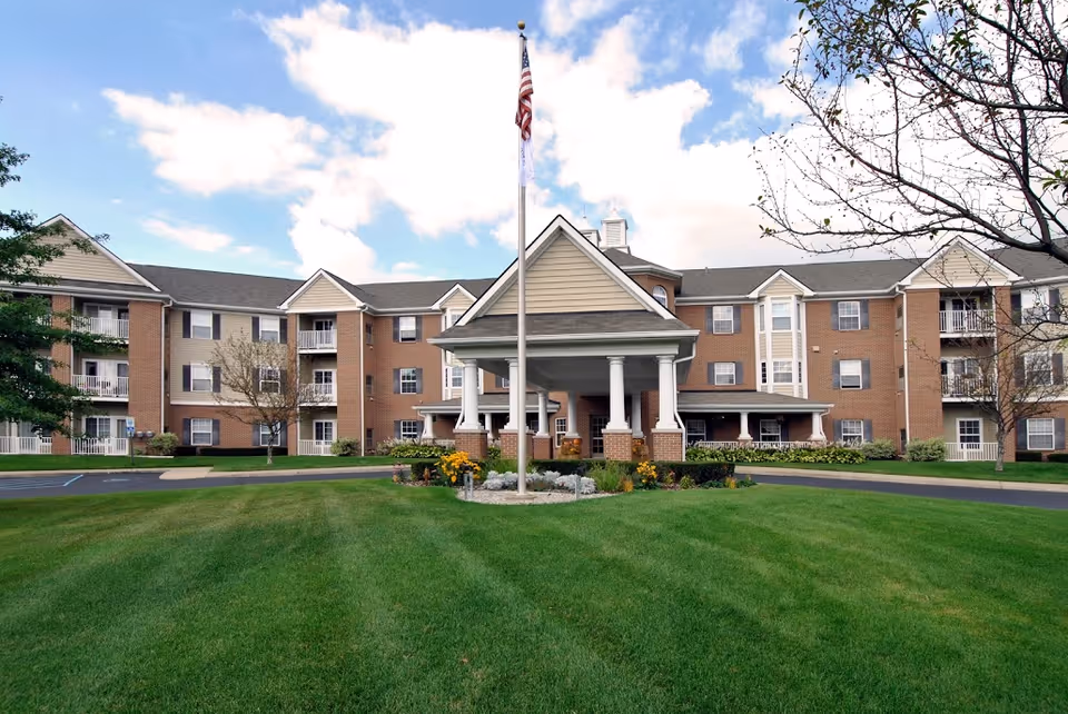 Brick senior living facility with a covered entrance, flagpole, and manicured lawn.