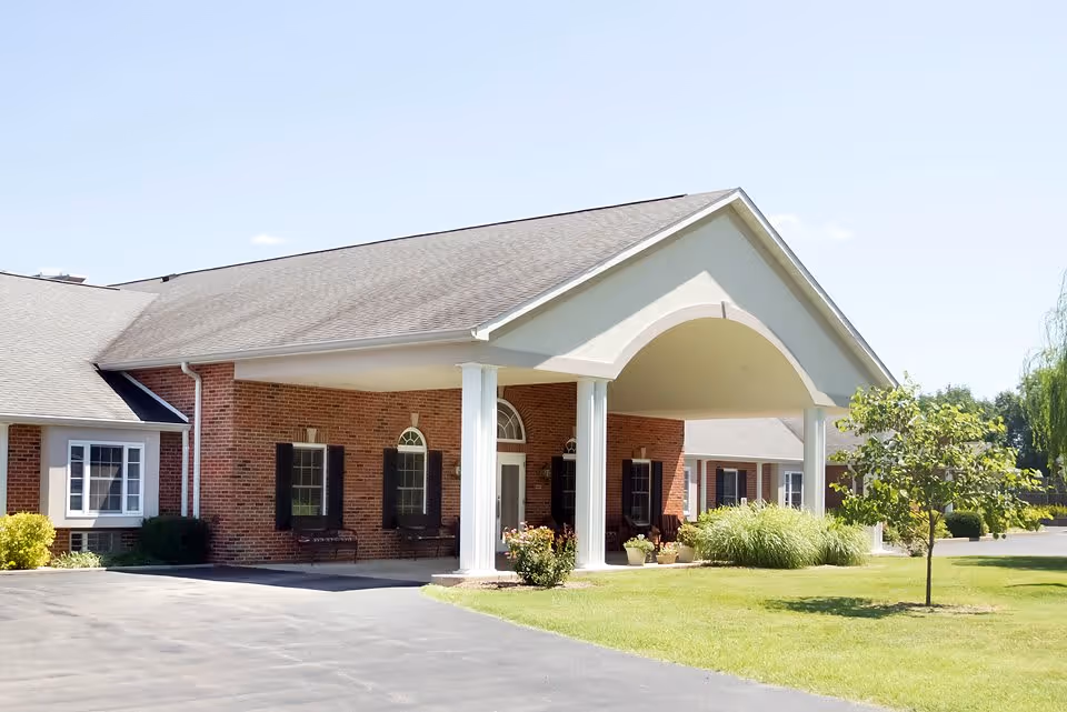 Front entrance of a brick senior living building with a covered portico, white columns, driveway and lawn.