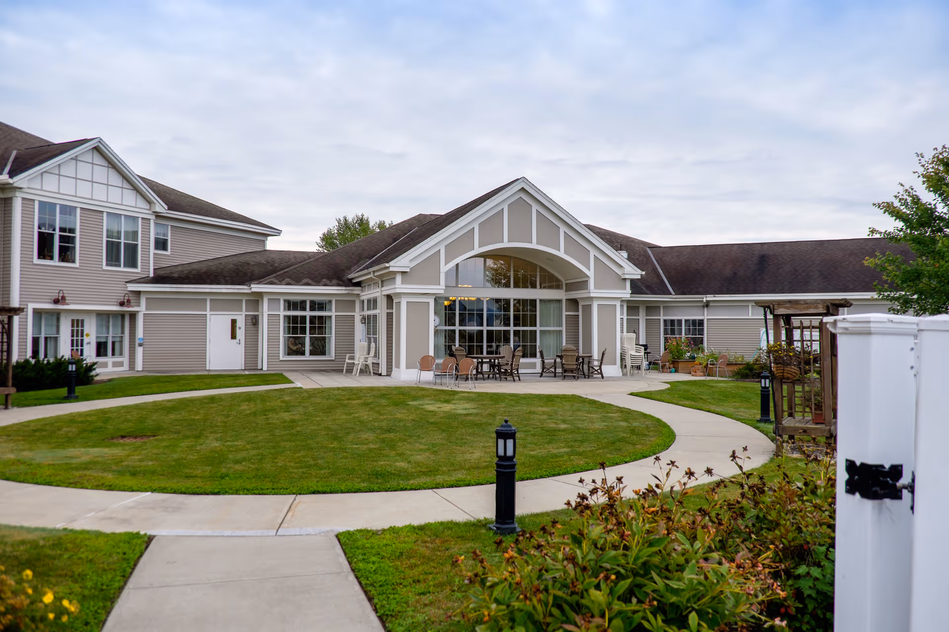 Exterior view of The Terrace at The Glen senior living facility showing a well-maintained lawn with a curved concrete walkway leading to a building entrance with large windows and outdoor seating under a covered patio.