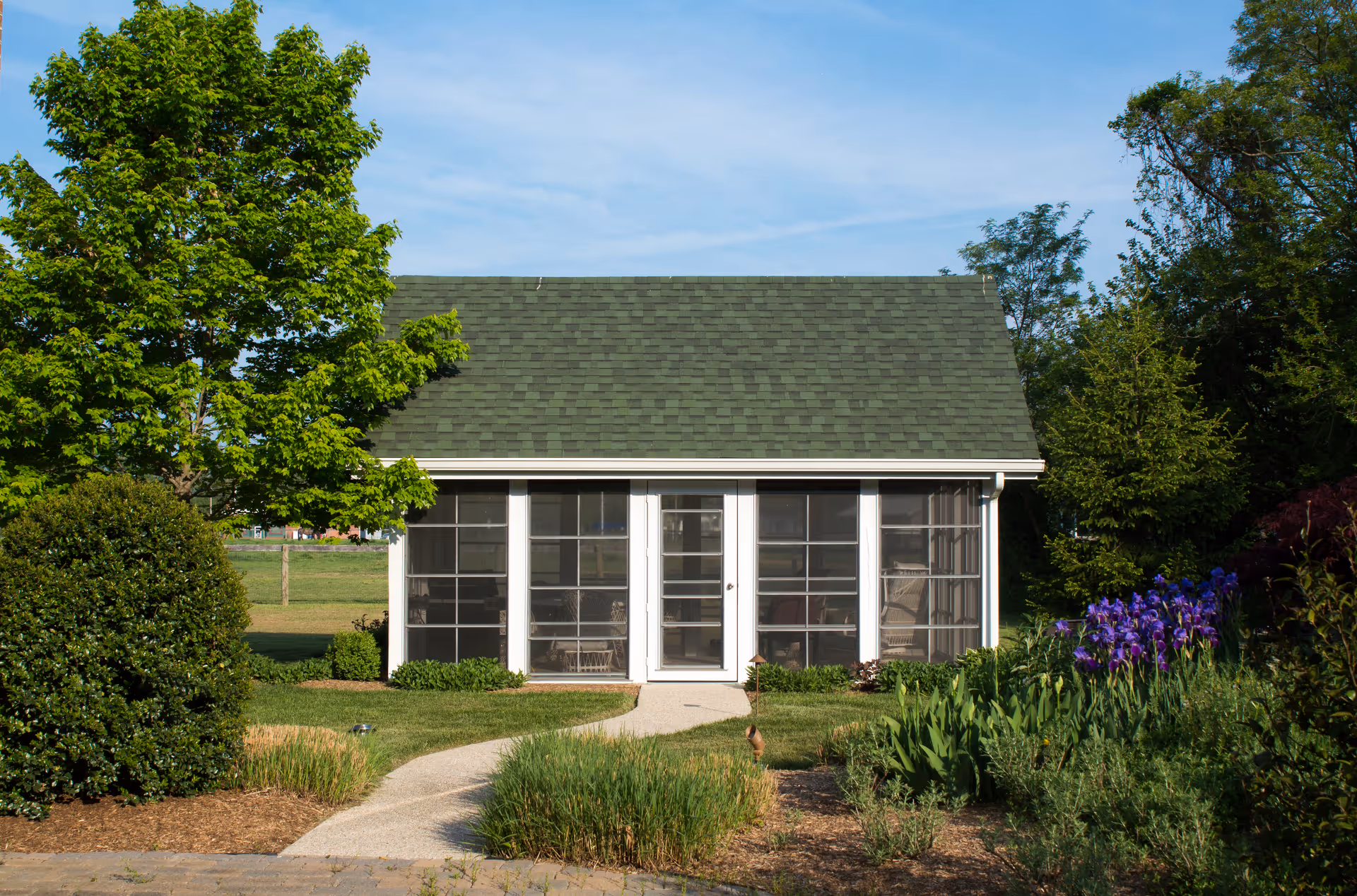 Small sunroom-style building with large glass doors, surrounded by shrubs and flowers.