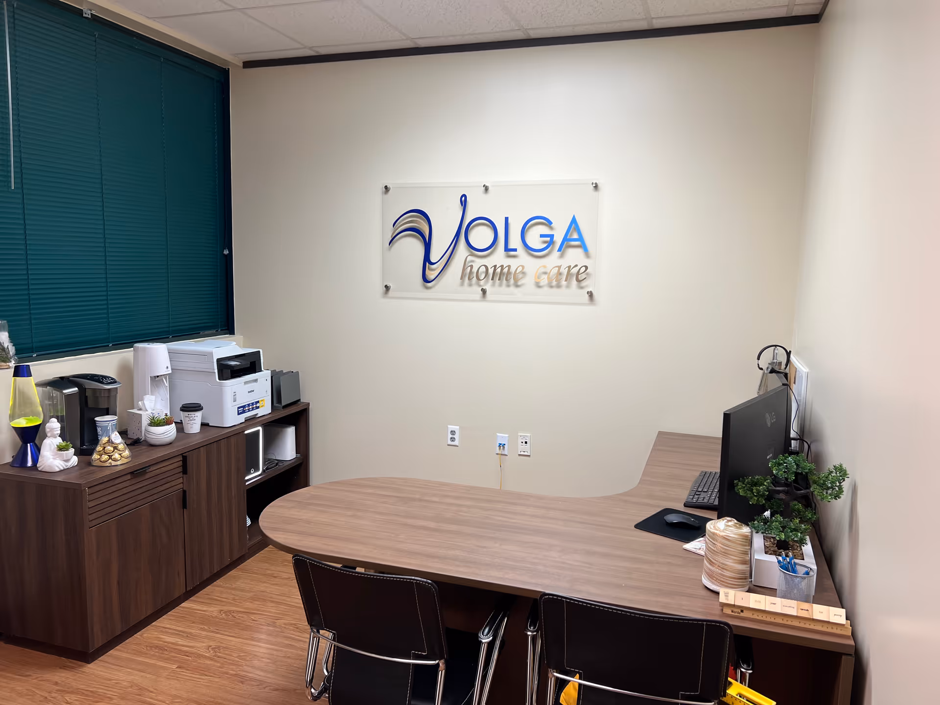 Office reception area with Volga Home Care sign, desk, computer, printer, and plants.