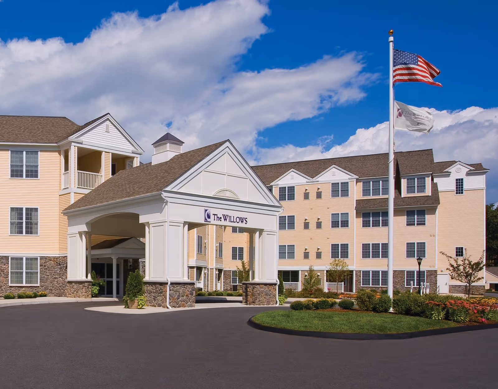 Exterior view of The Willows Retirement Community building with a covered entrance, landscaped grounds, and two flags on a flagpole against a partly cloudy blue sky.