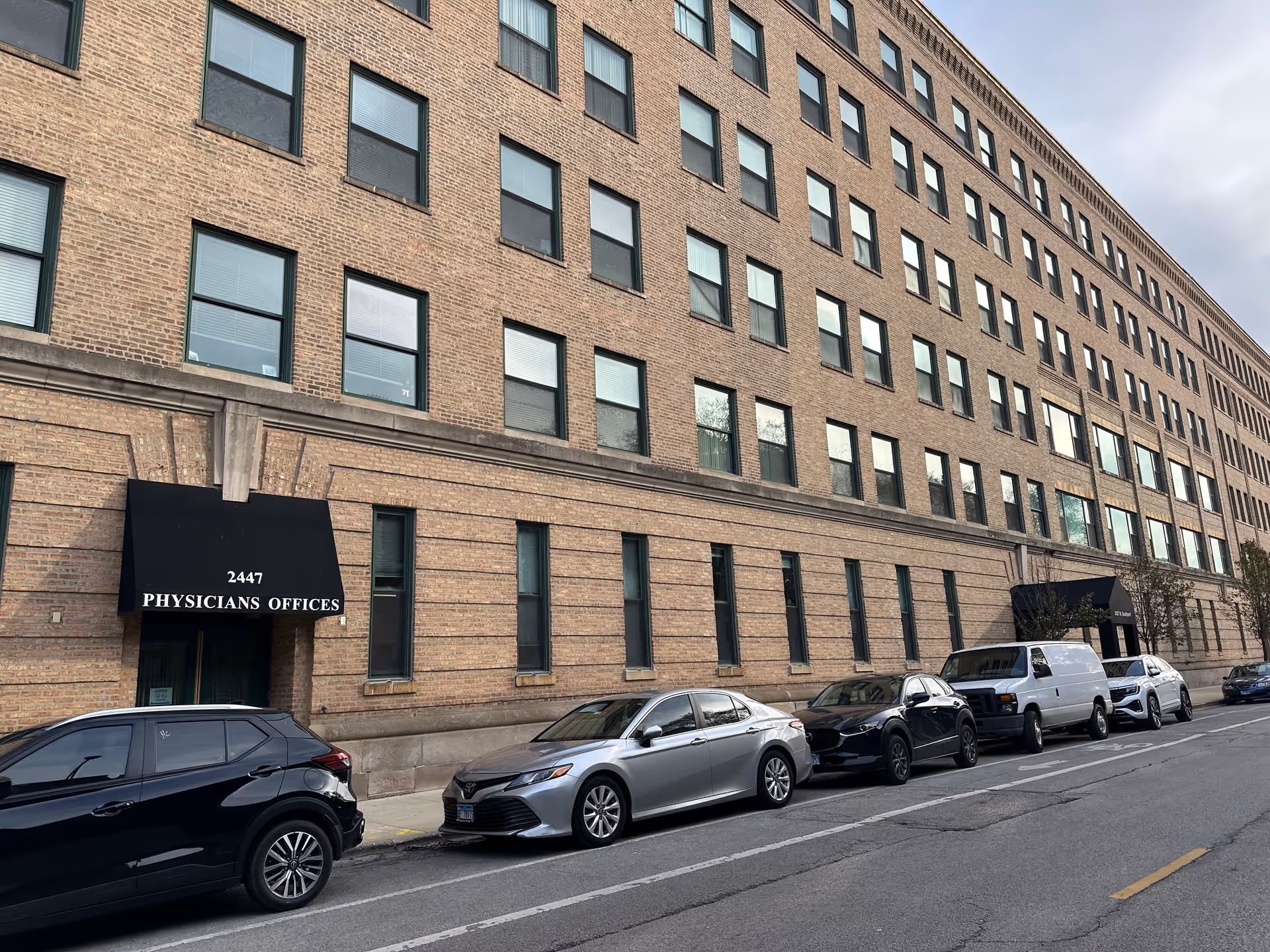 Exterior of a multi-story brick building with many windows, awnings marked "2447 PHYSICIANS OFFICES", and cars parked along the street.