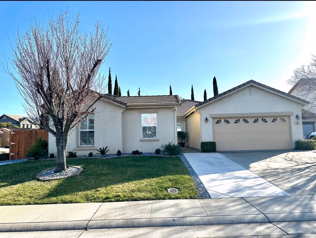 Beige, single-story house with a two-car garage and leafless tree in front yard.