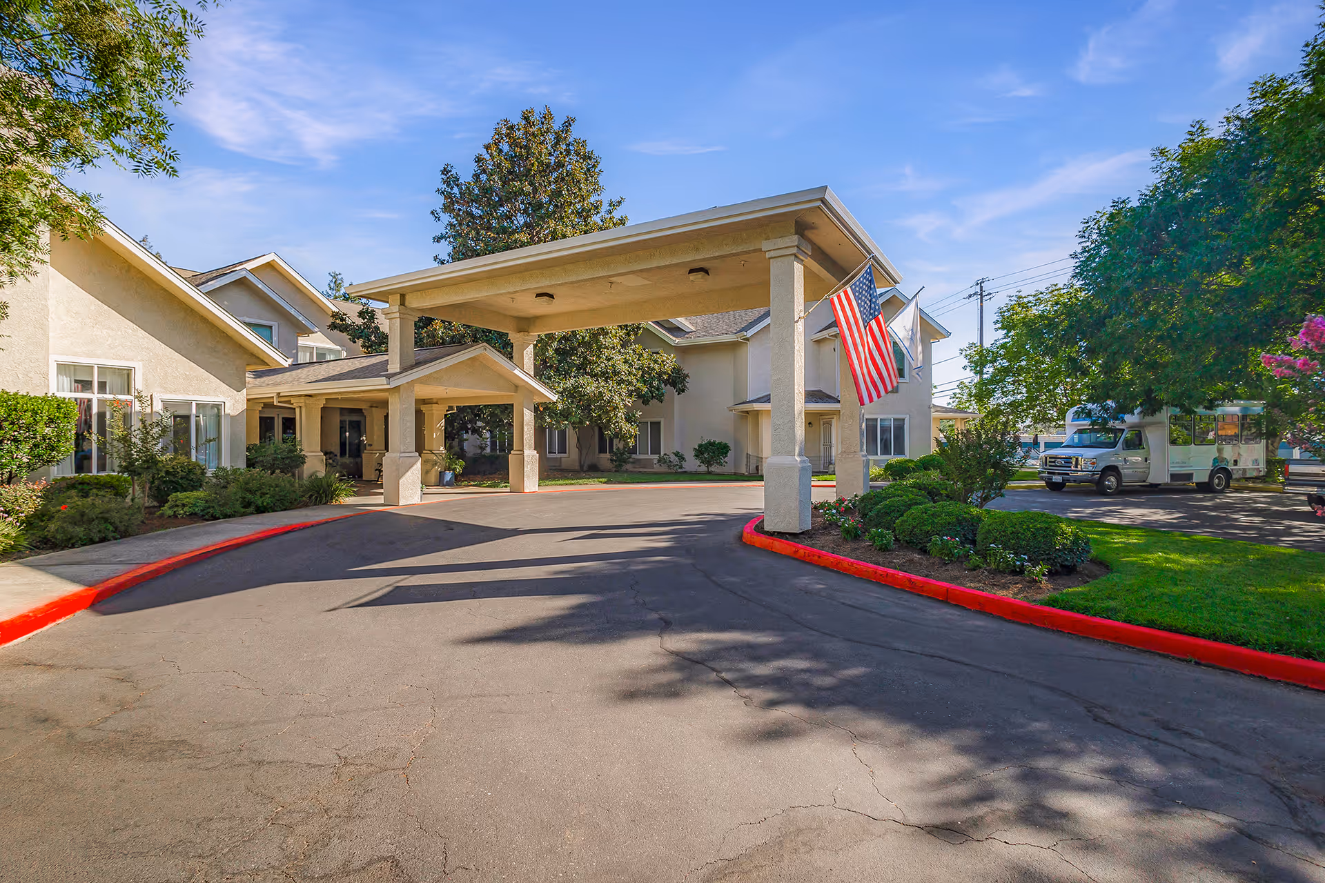 Exterior view of Marbella Chico facility entrance with a covered drop-off area supported by columns, American flags hanging, surrounding greenery, and a parked vehicle on the right side under clear blue sky.