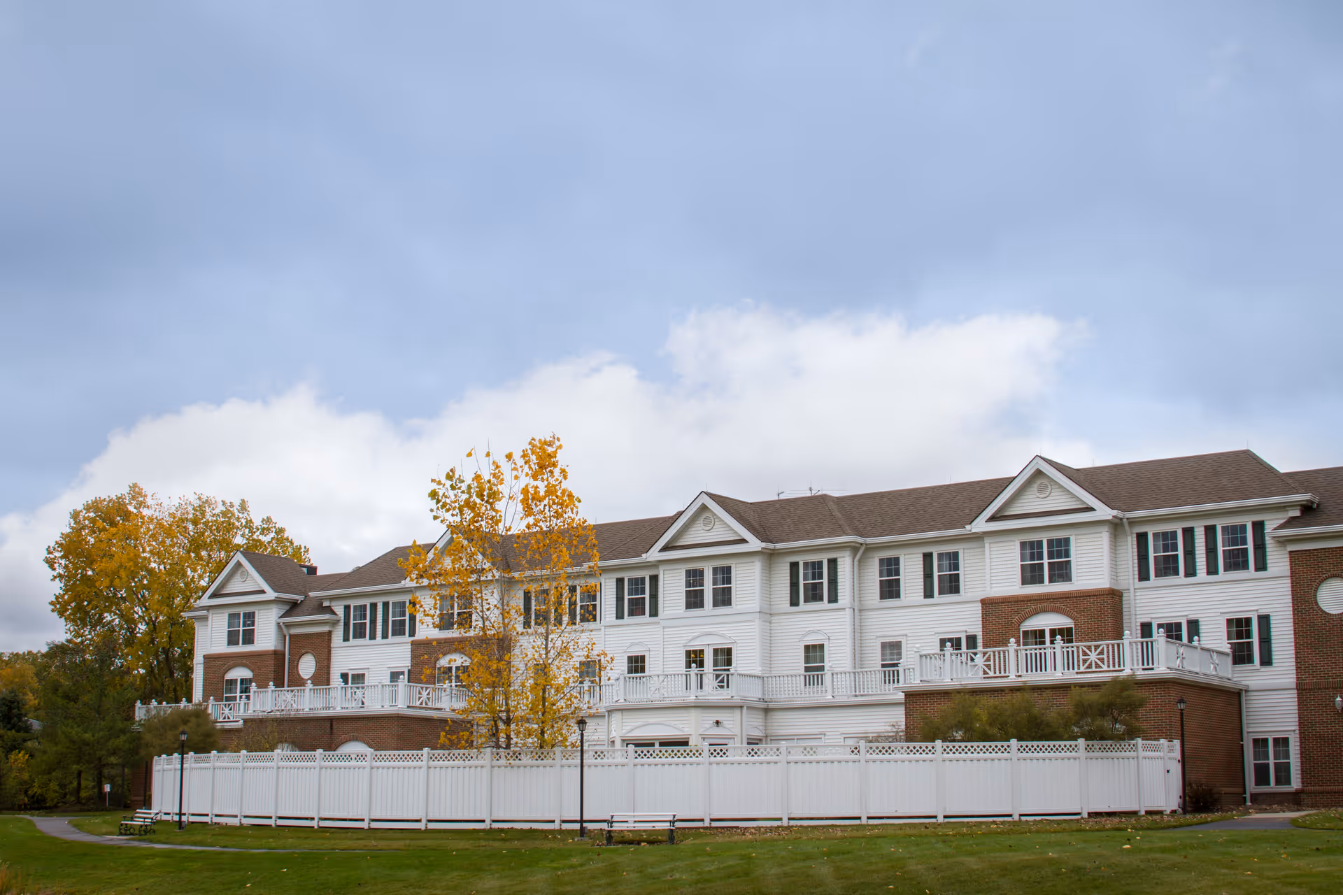 White multi-story senior living building with brick accents, fenced lawn, and autumn trees.