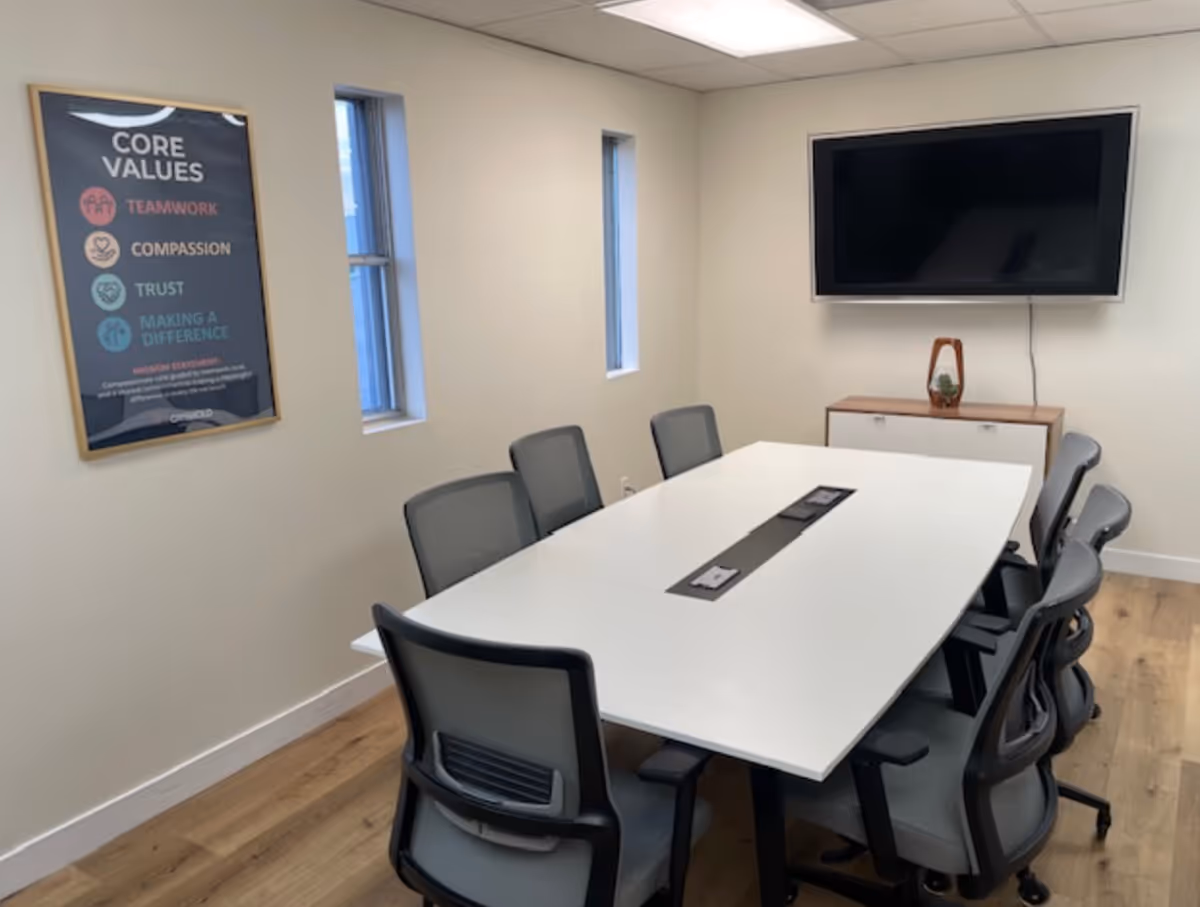A conference room with a long white table, chairs, a wall poster, and a mounted TV.