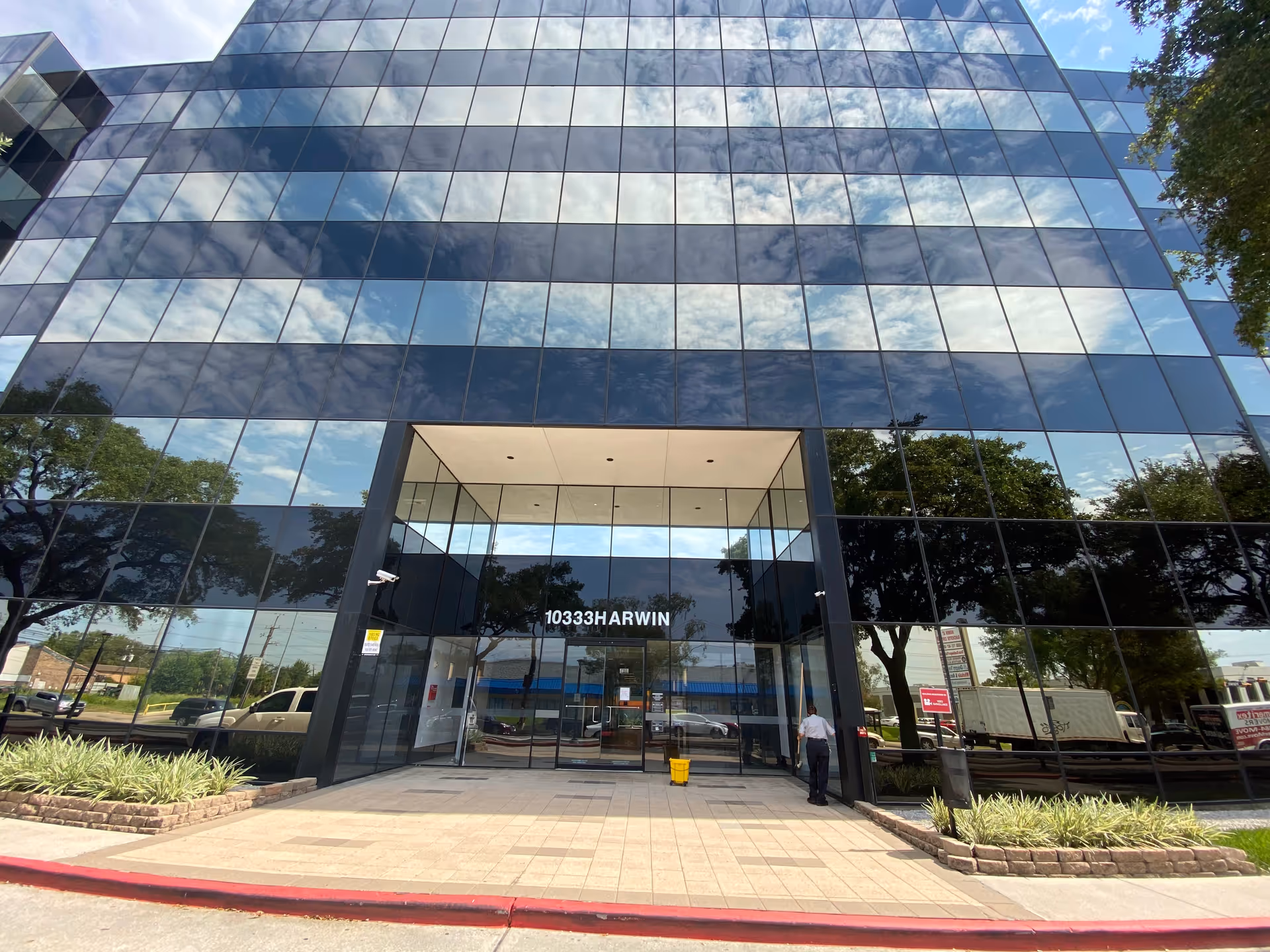 Glass-front office building entrance reflecting trees and sky with a sidewalk and a person